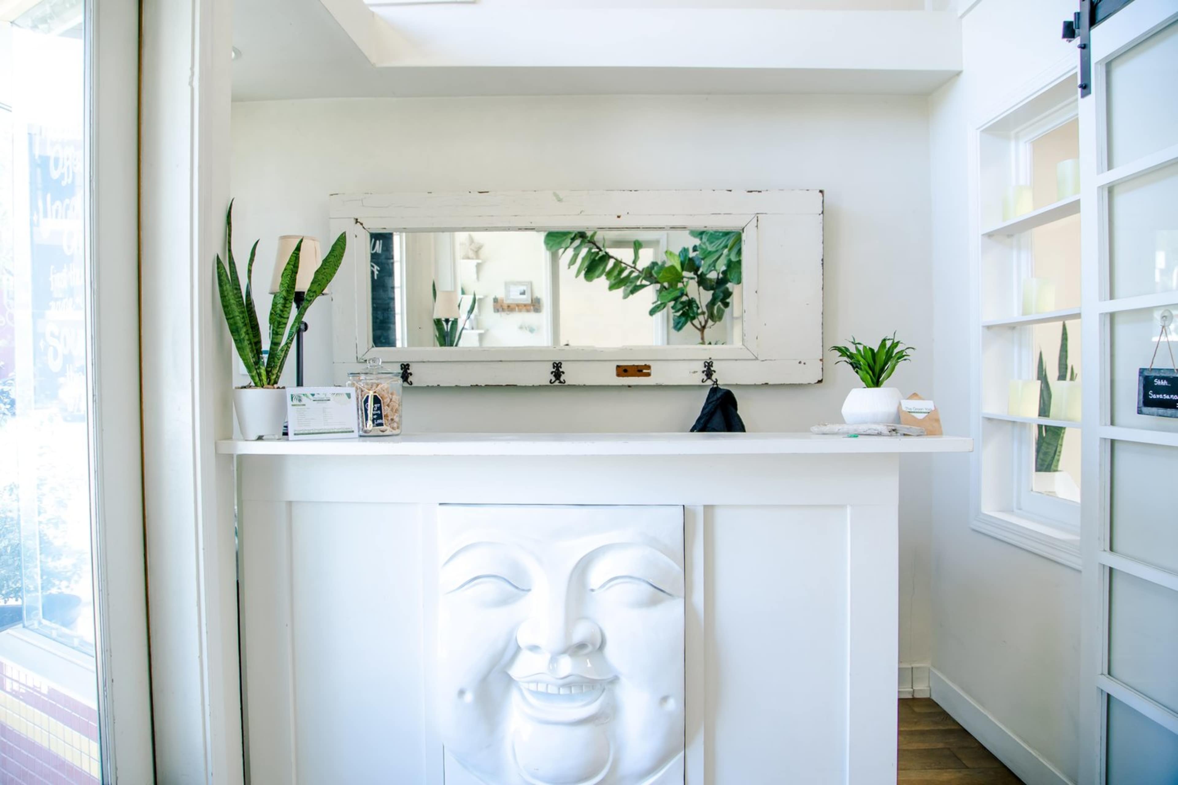 A reception area featuring a white counter with a smiling face relief and a large mirror above it, flanked by potted plants.