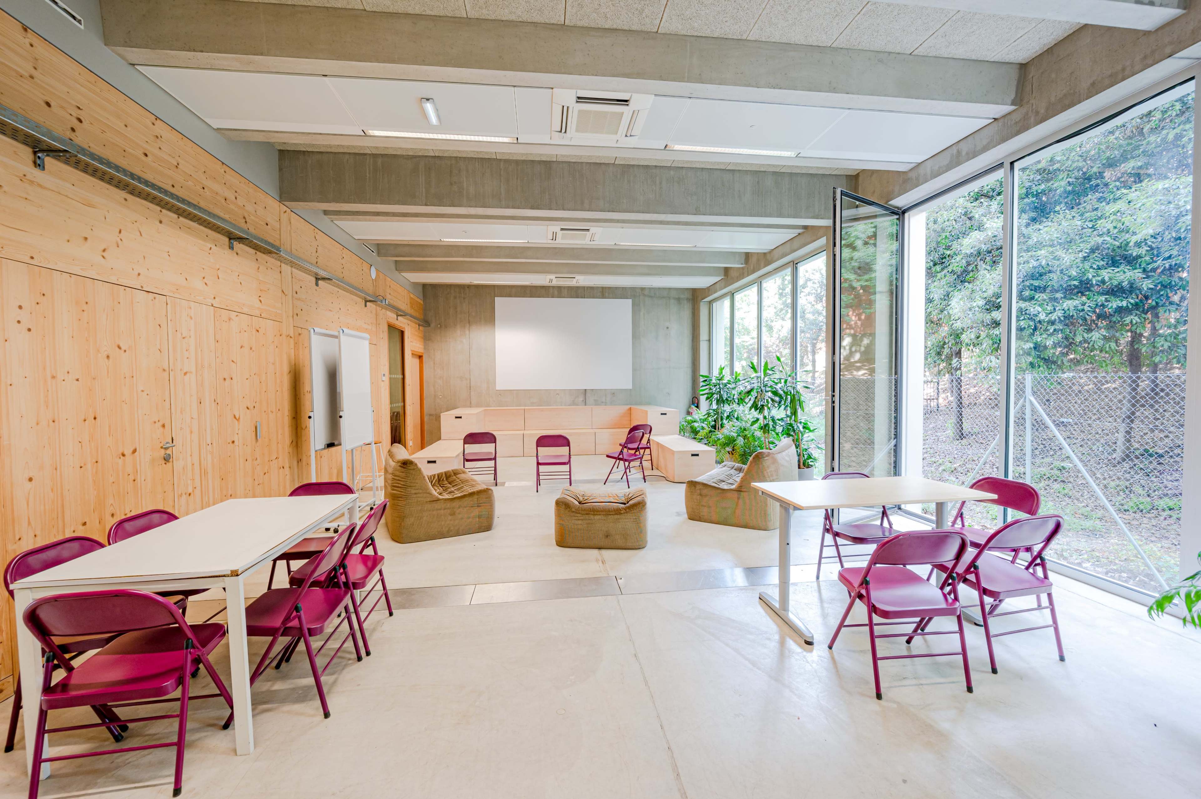 A modern classroom with several tables and chairs arranged around a central seating area, surrounded by large windows and greenery outside.