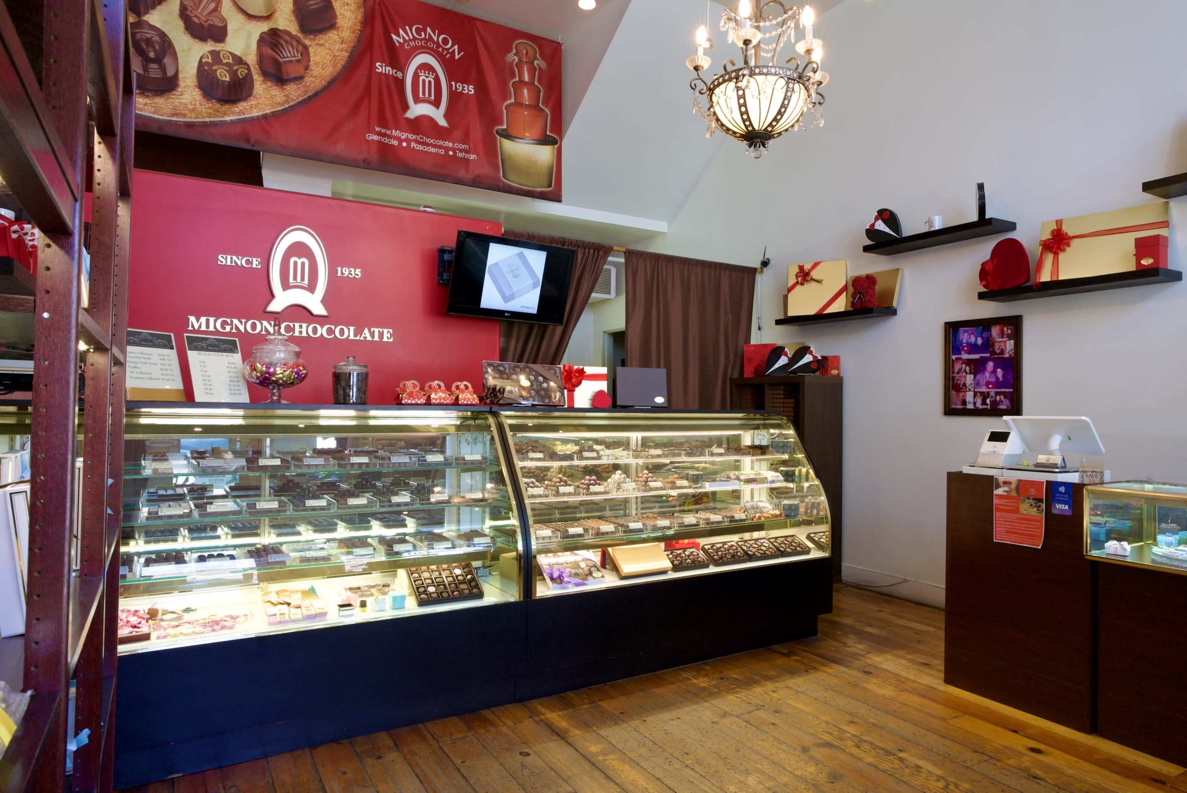 The interior of a chocolate shop features glass display cases filled with various chocolates, a prominent sign reading "Mignon Chocolate," and elegant decor including a chandelier.