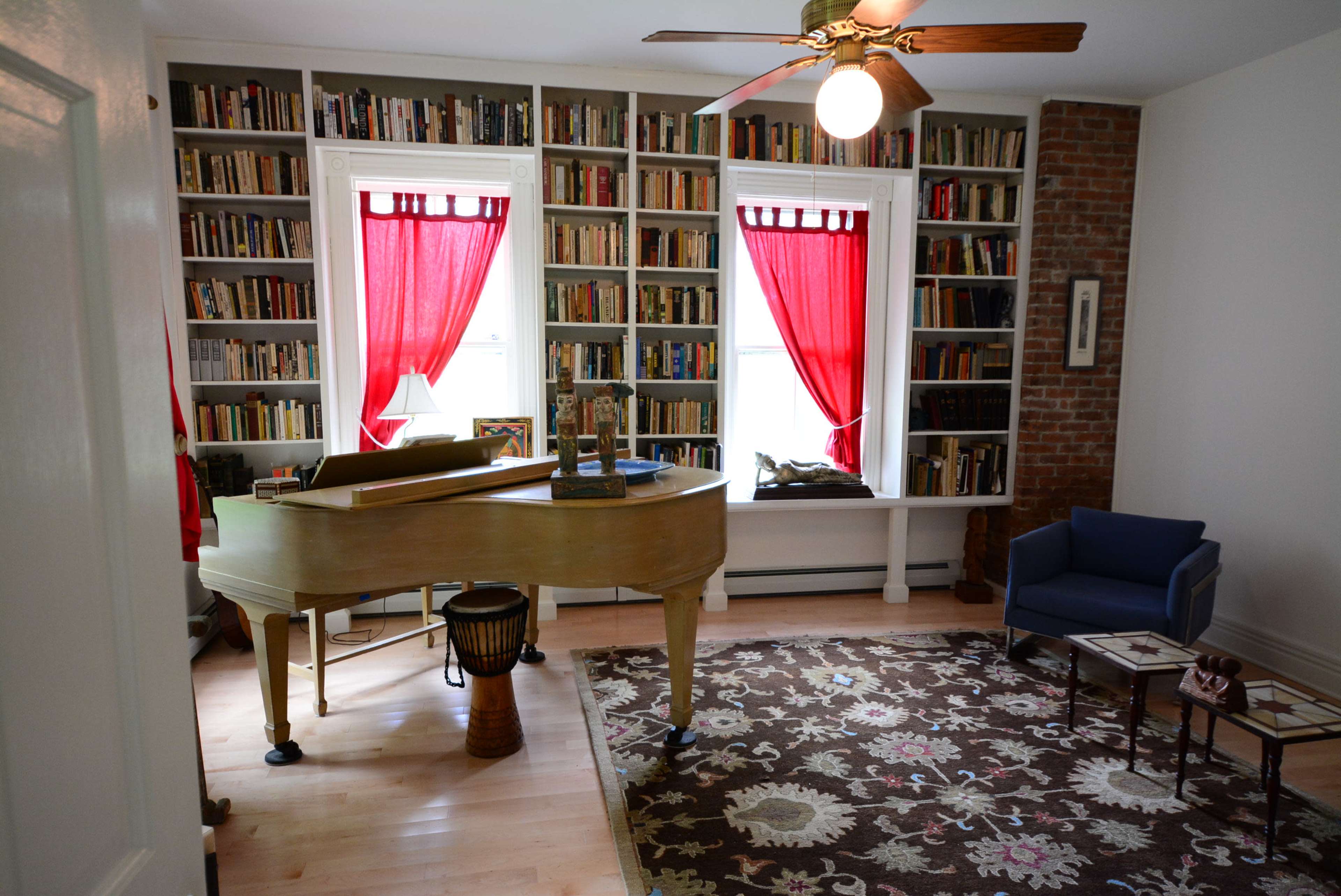 The image shows a room with a grand piano, a blue armchair, and bookshelves filled with books, accented by red curtains at the windows.