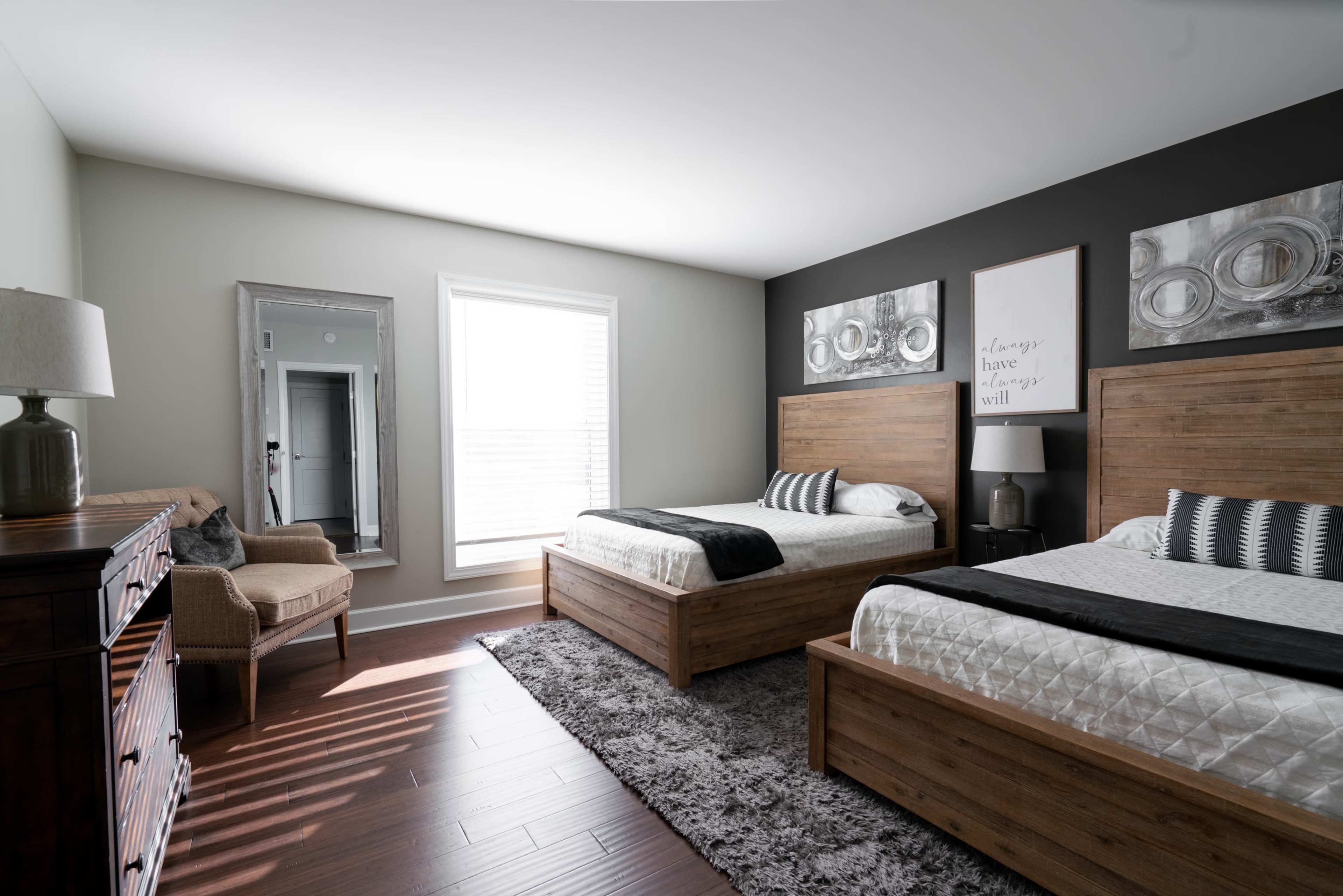 A well-lit bedroom featuring two wooden beds, a gray area rug, a mirror, a small armchair, and wall art above the beds.