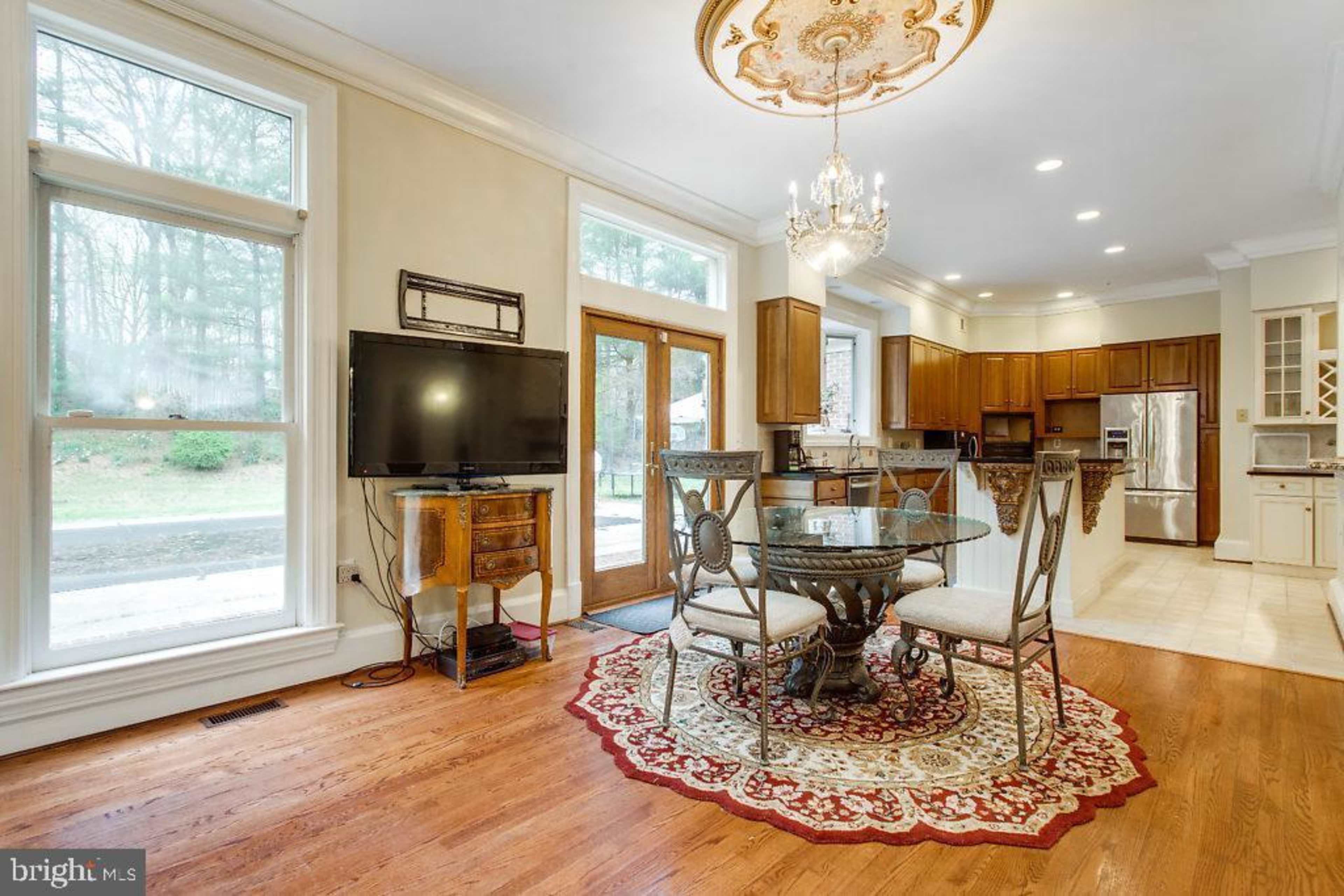 A dining area with a round table and four chairs, featuring a chandelier overhead and a television on a stand, with large windows allowing light to enter from outside.