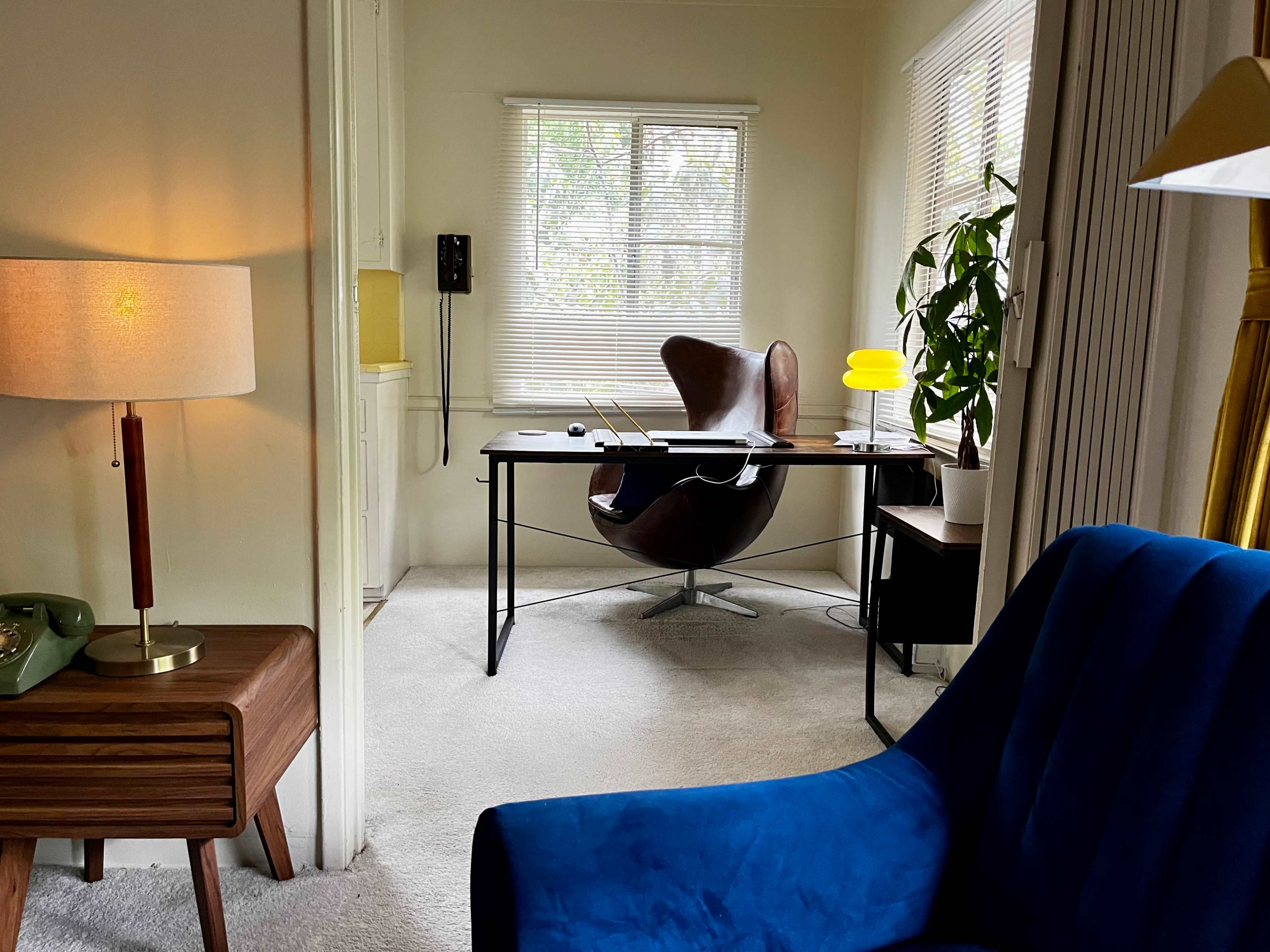 A modern workspace features a sleek desk with a chair, a lamp, a plant, and large windows providing natural light.