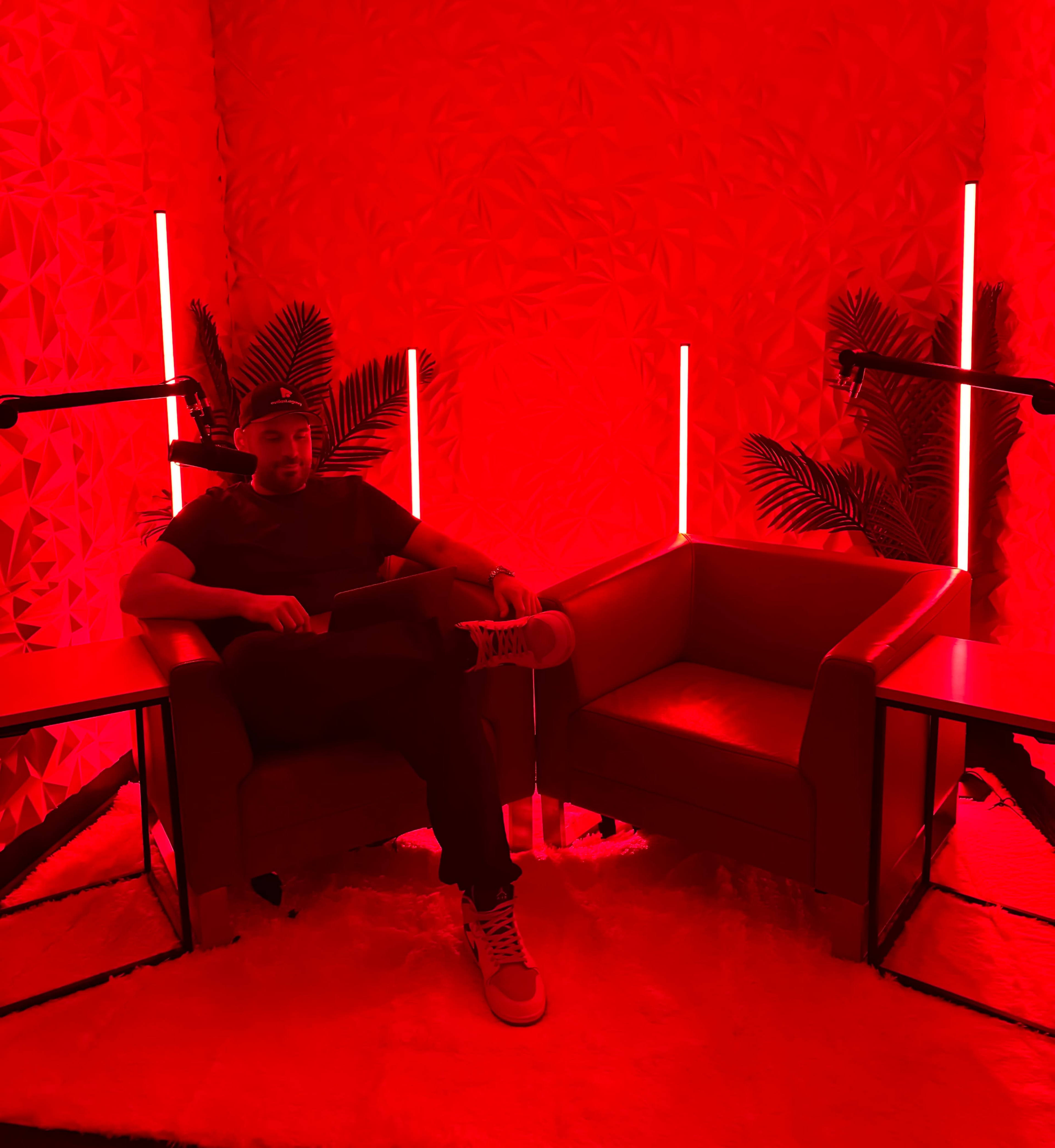 A person sits between two chairs in a red-lit room with plant decorations and microphones.