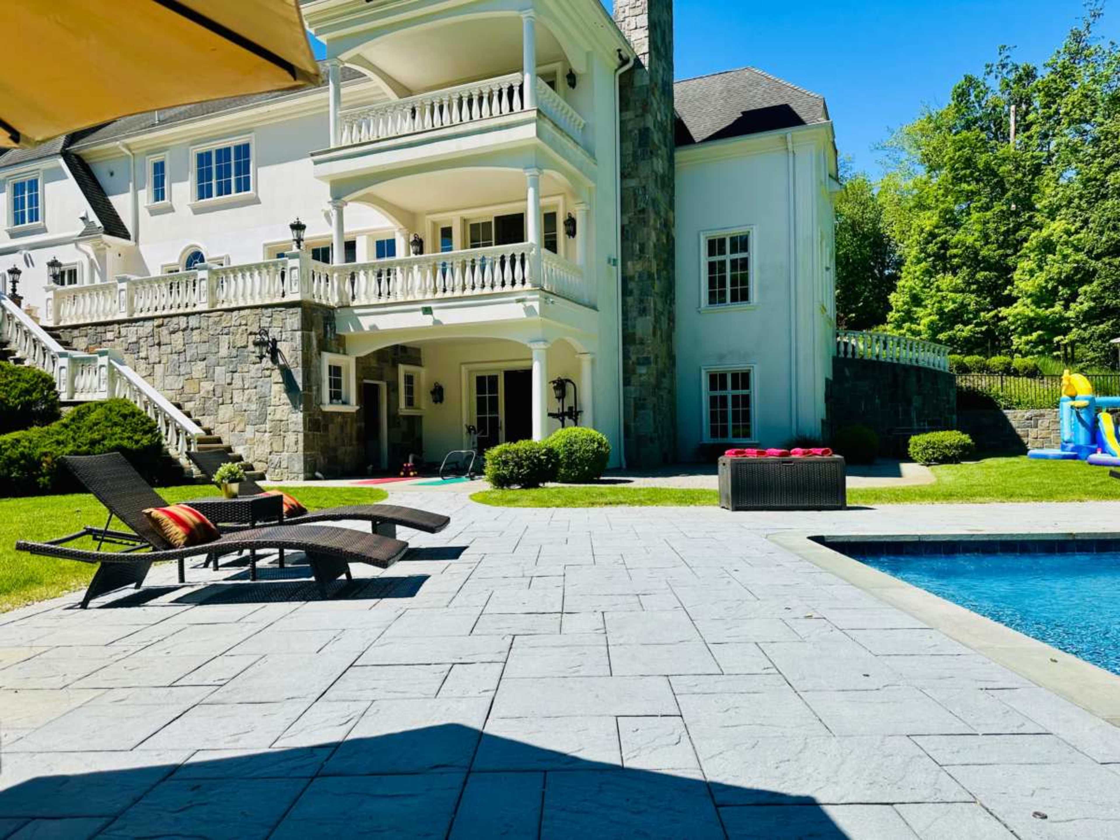 The image shows a spacious backyard with a stone patio, a swimming pool, and a large house featuring multiple balconies.