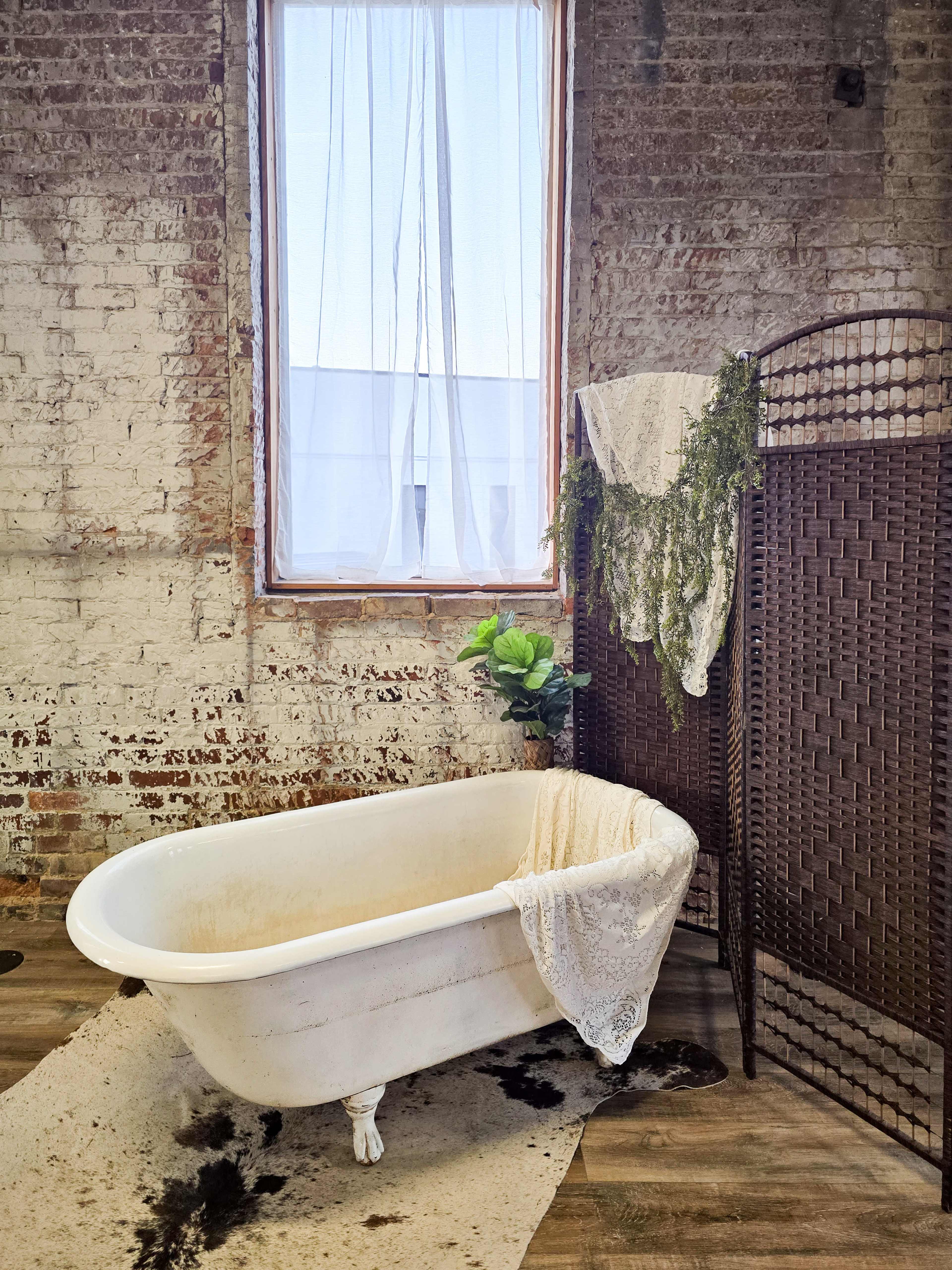 The image shows a vintage clawfoot bathtub placed on a wooden floor, next to a woven screen and decorated with plants and a white lace cloth.