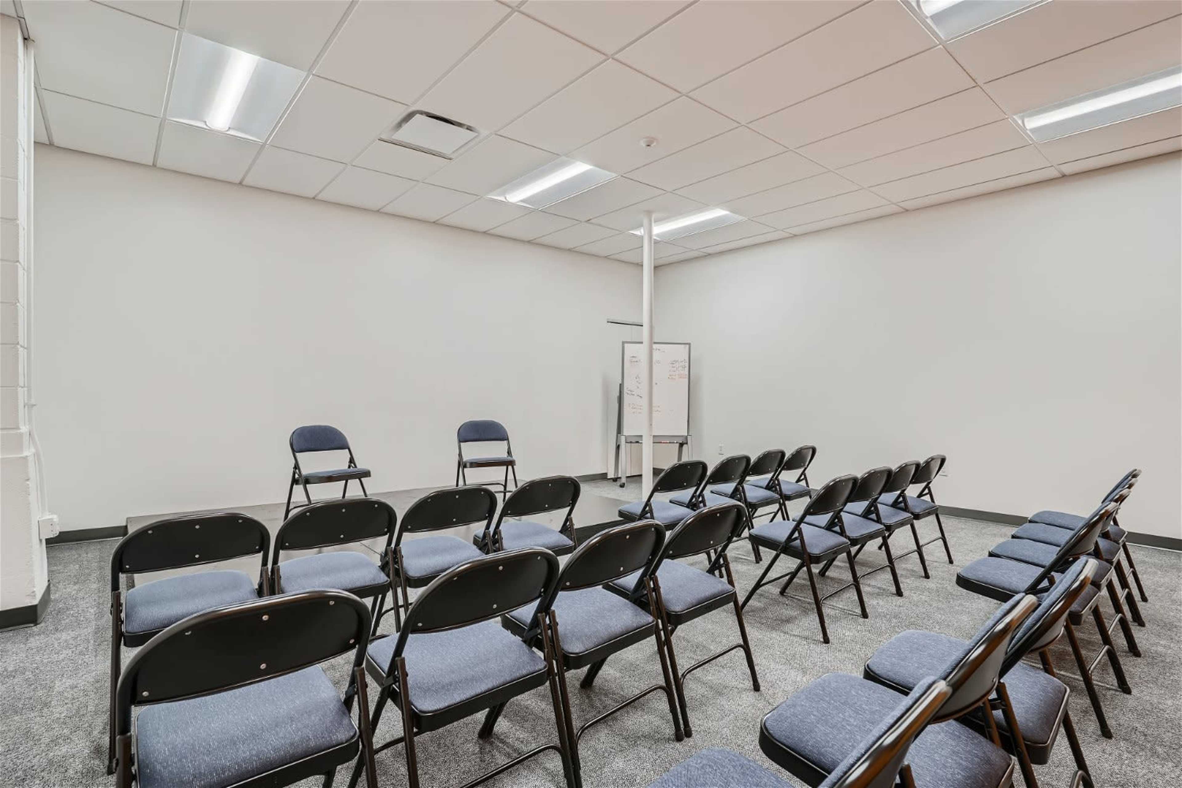 A sparsely furnished meeting room features rows of black folding chairs facing a small area with two empty chairs and a whiteboard.