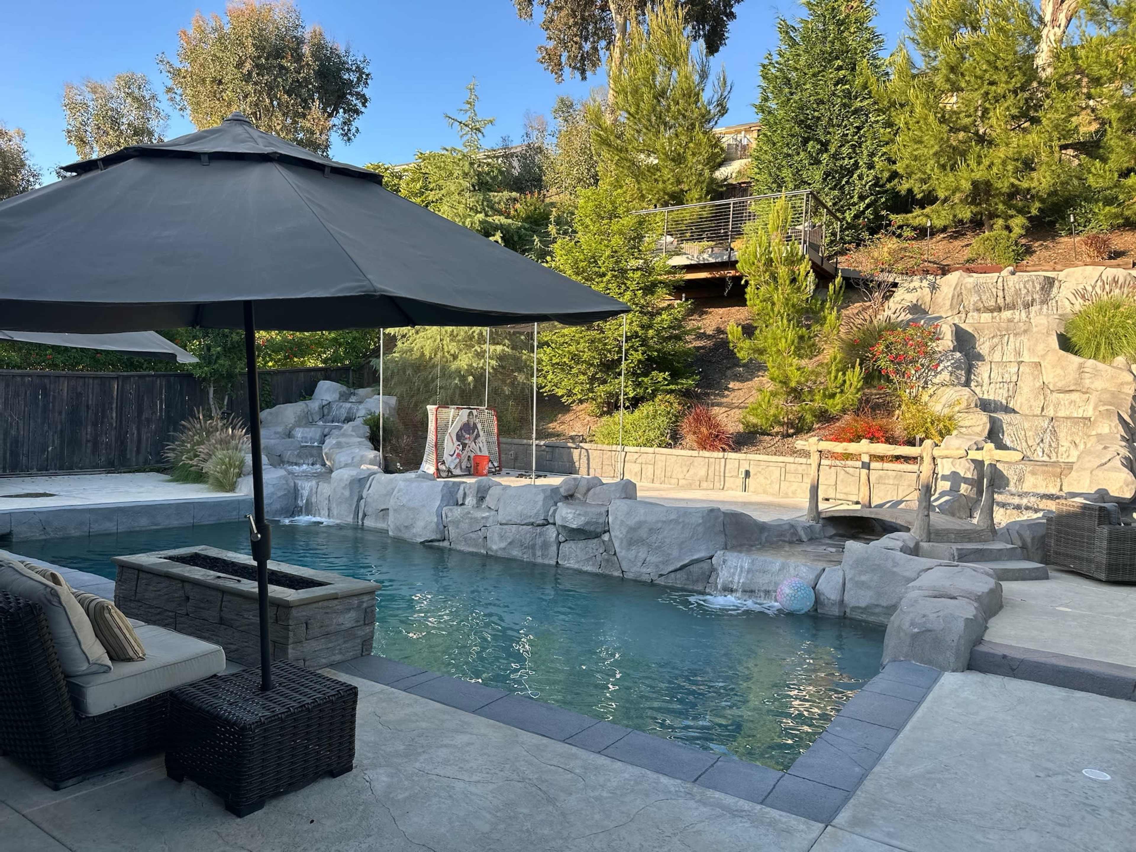 A backyard pool area featuring a rock waterfall, a black umbrella over patio furniture, and landscaped greenery in the background.