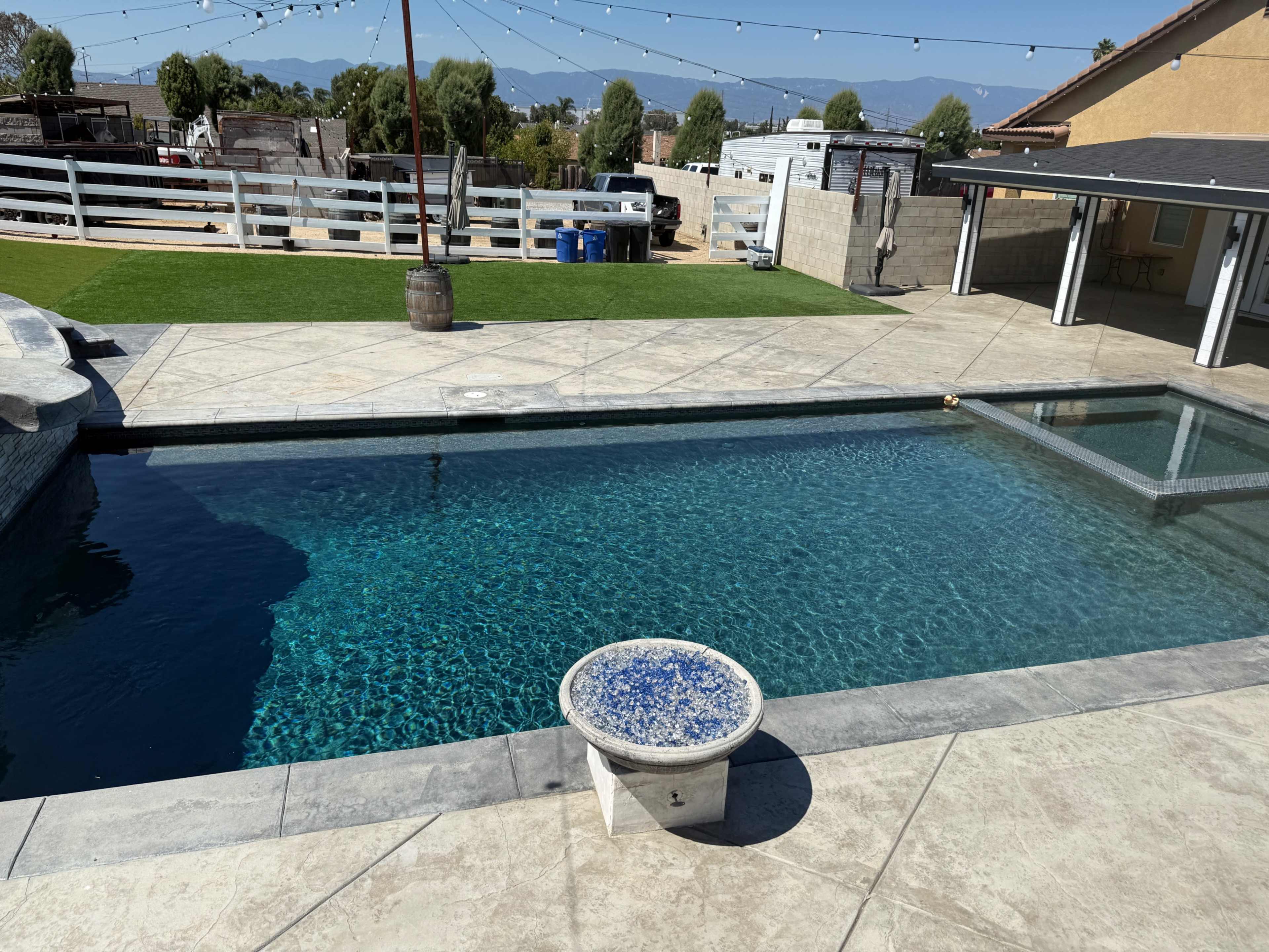 The image shows a clear swimming pool with a stone deck, surrounded by a lawn and a fenced area, under a blue sky.