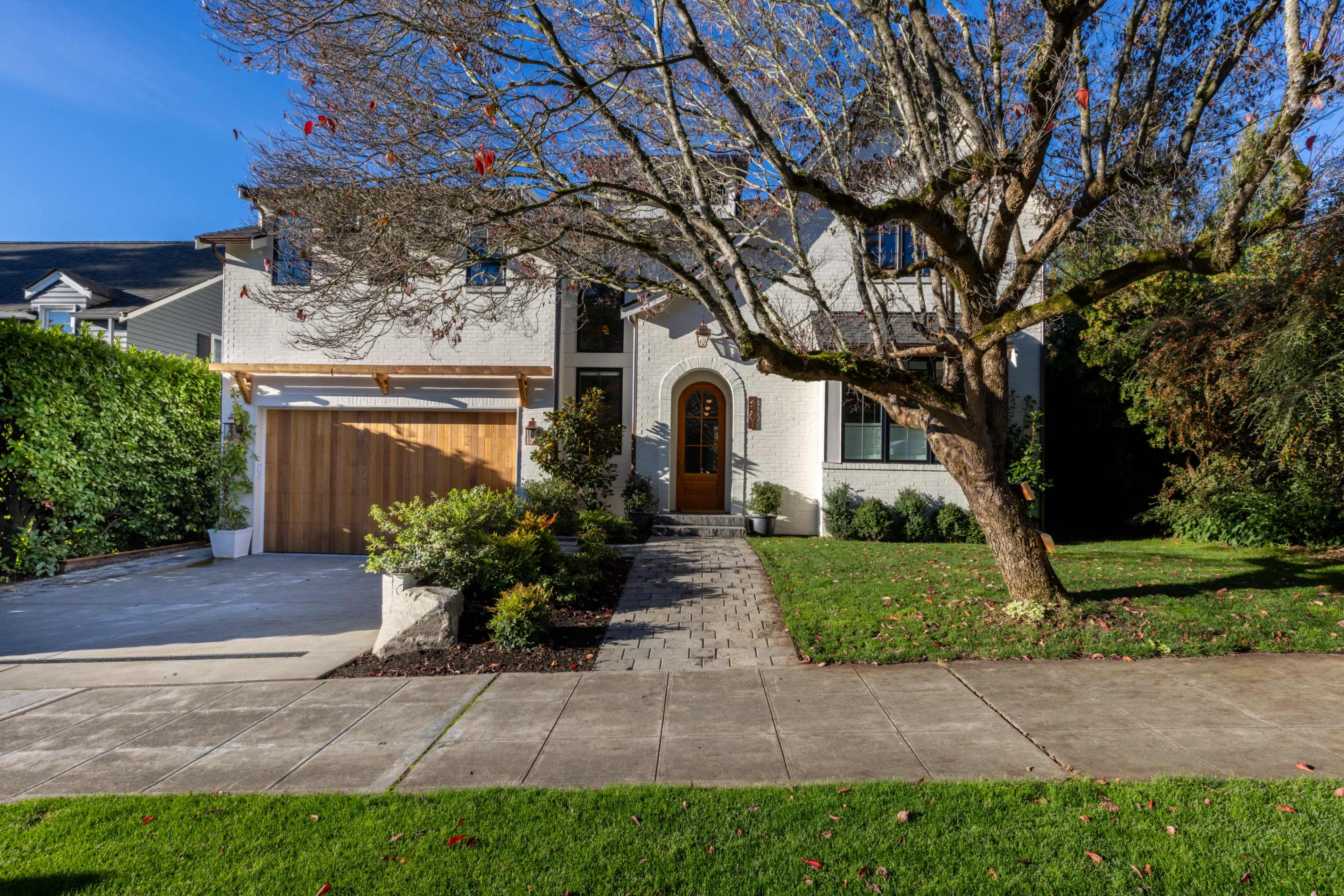 The image shows a two-story house with a landscaped front yard, a driveway leading to a wooden garage door, and a large tree in the foreground.