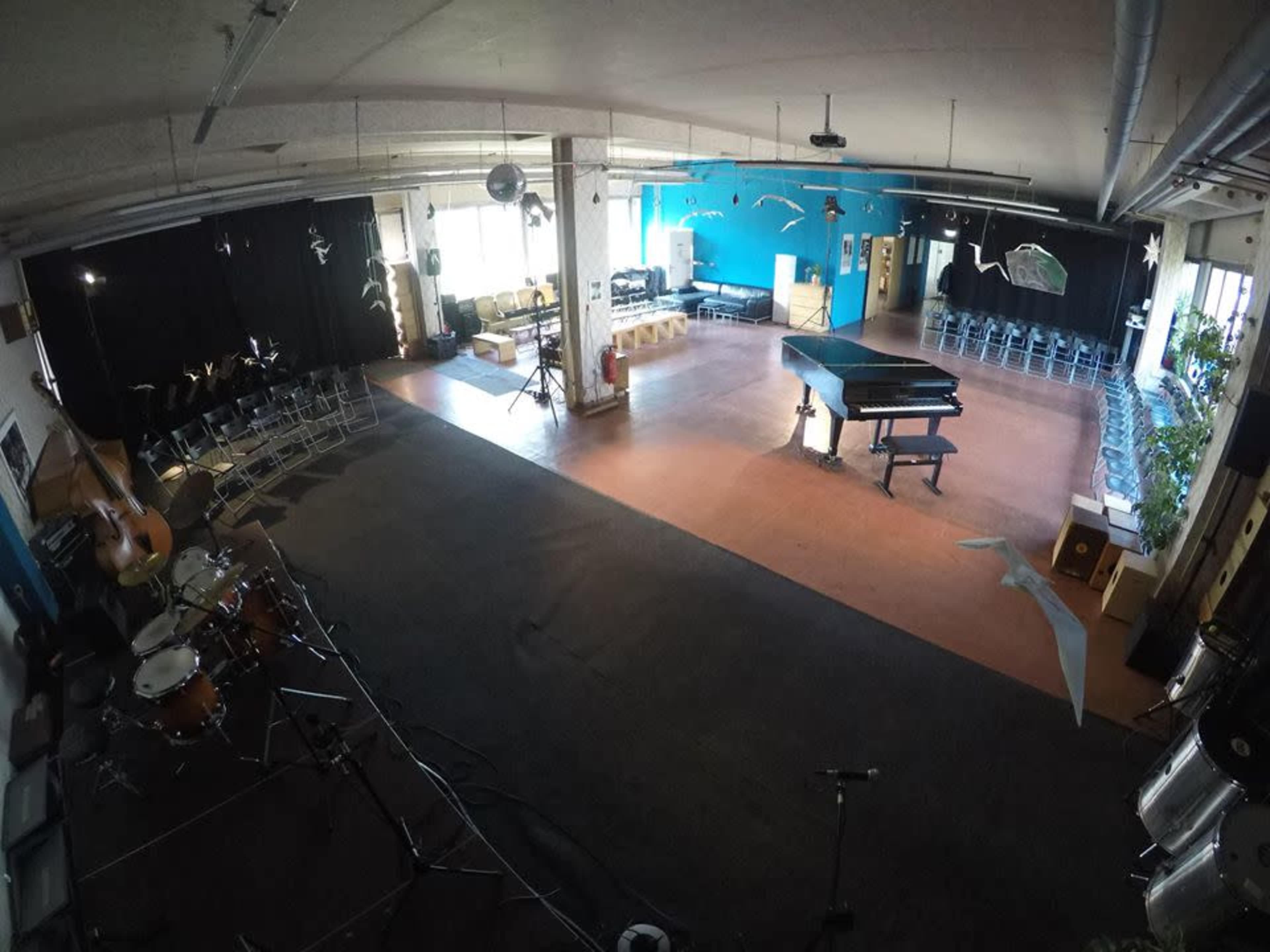 The image shows a large, open space featuring a grand piano, musical instruments, chairs arranged for an audience, and a backdrop of blue walls with decorations.
