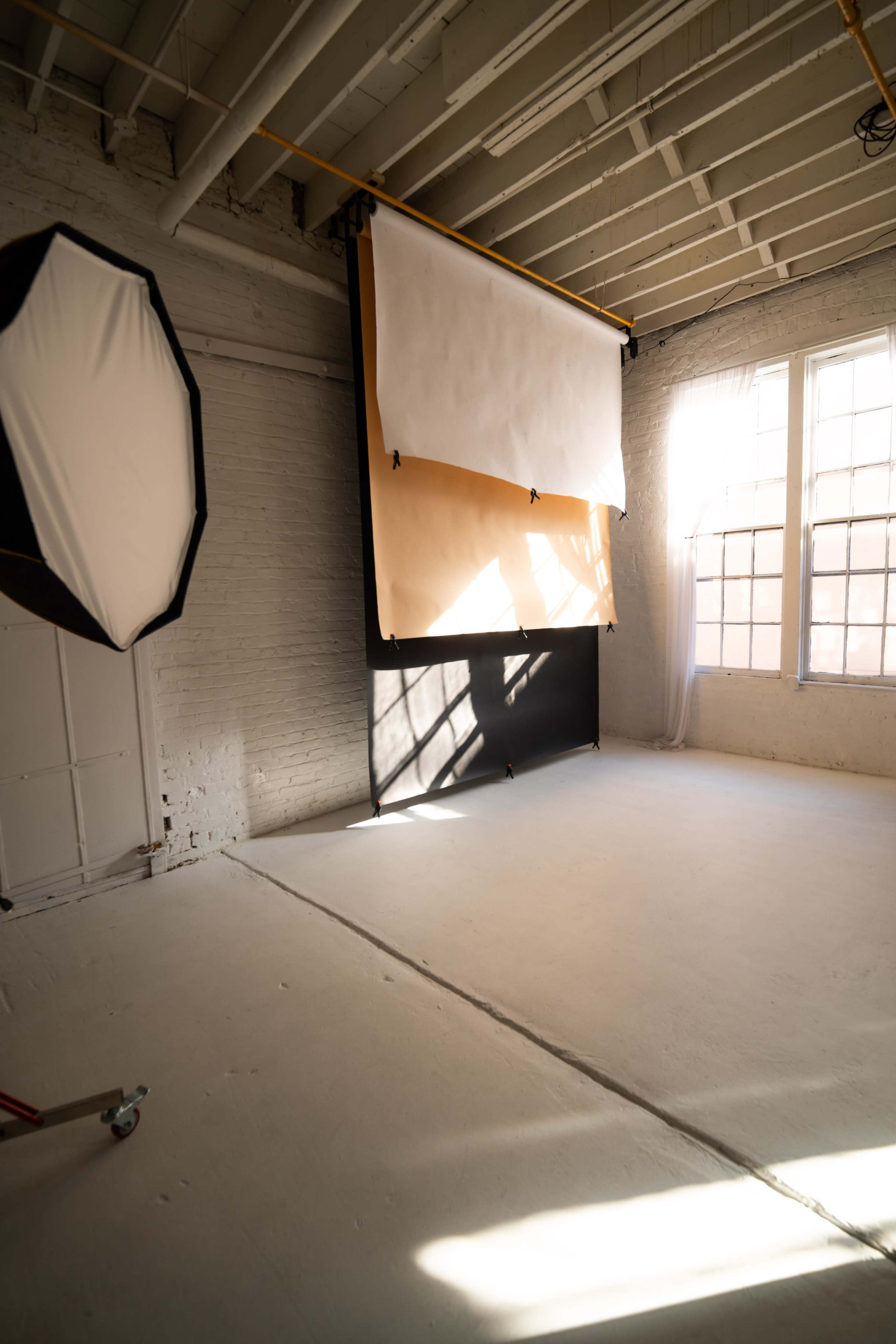 The image shows a bright, empty photography studio with a paper backdrop, large softbox lighting, and windows allowing natural light.
