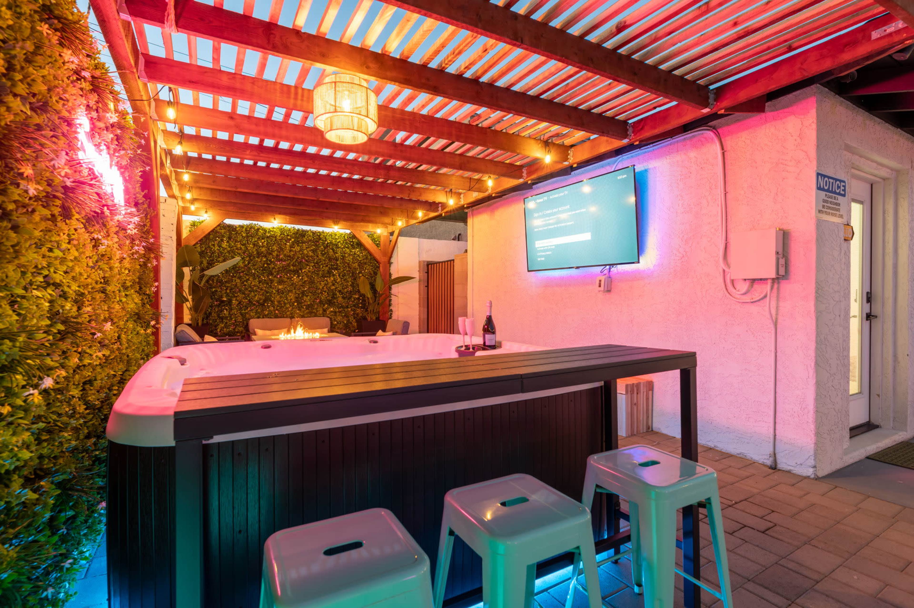 The scene features a well-lit outdoor area with a hot tub, a bar with stools, and a television mounted on the wall, surrounded by decorative plants and ambient lighting.