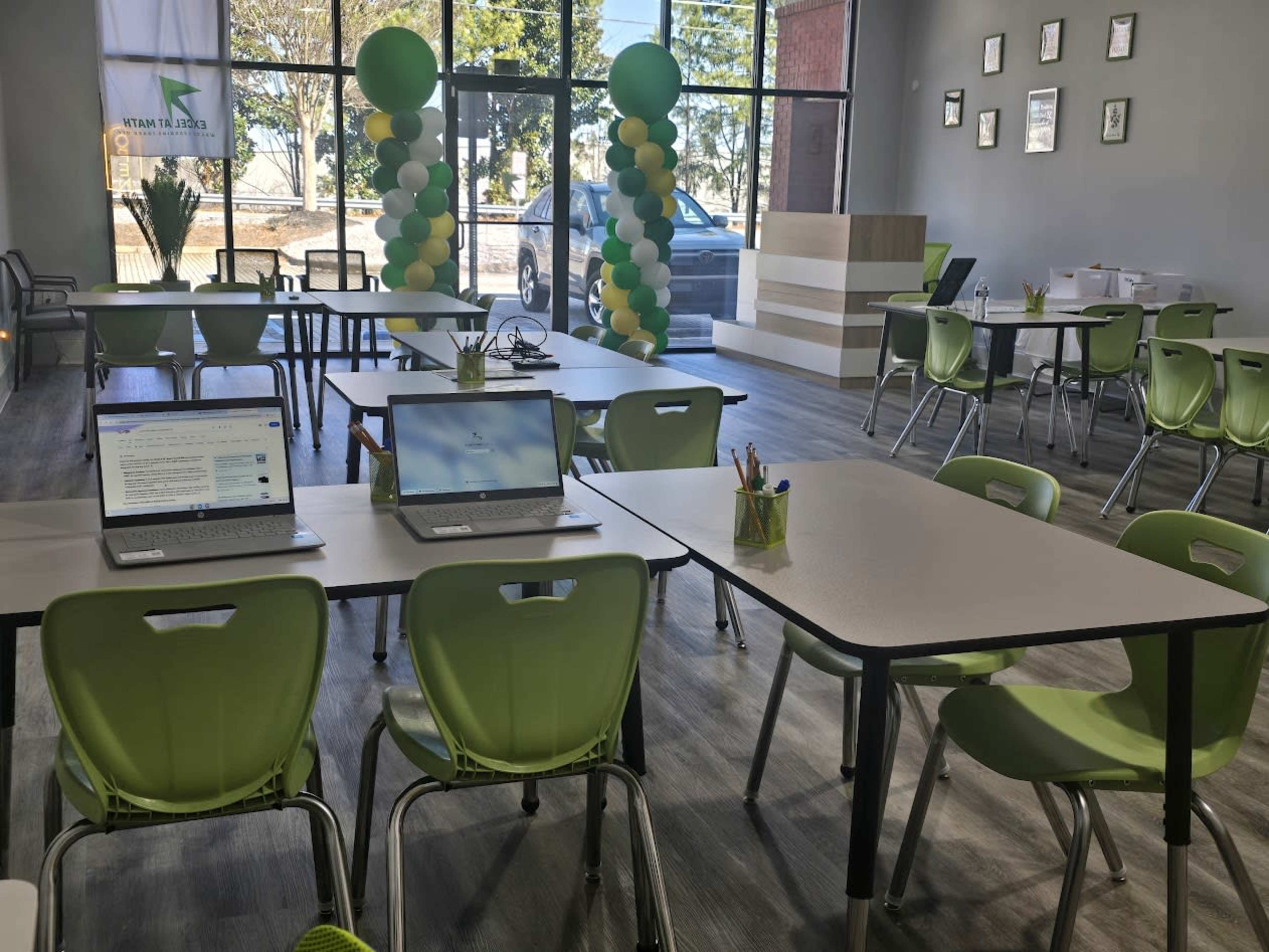 The image shows a modern classroom or study space featuring multiple tables with green chairs, two laptops opened on the tables, and a decorative balloon arch in the background.