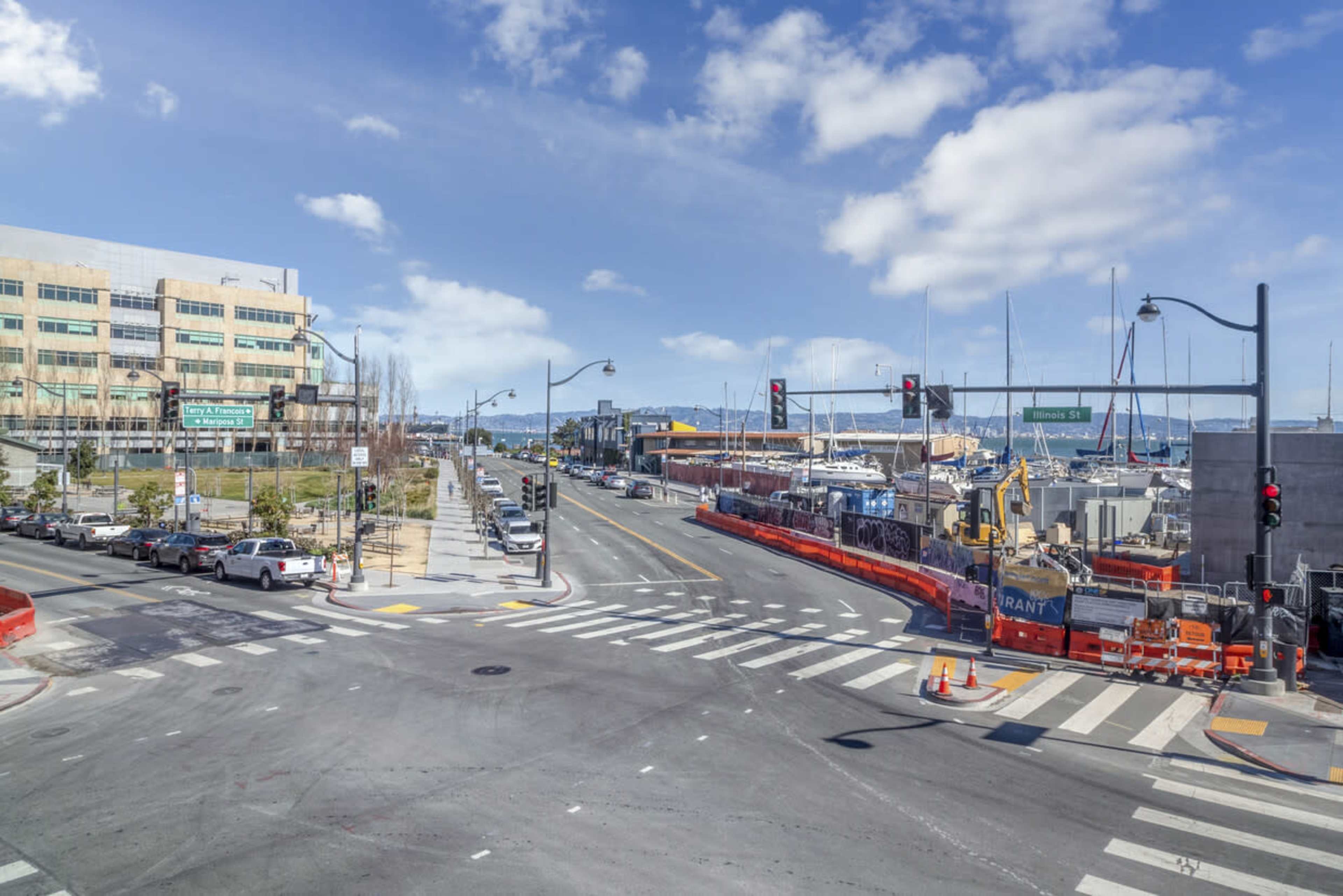 A street intersection near a harbor with construction barriers, parked cars, and sailboats in the water.