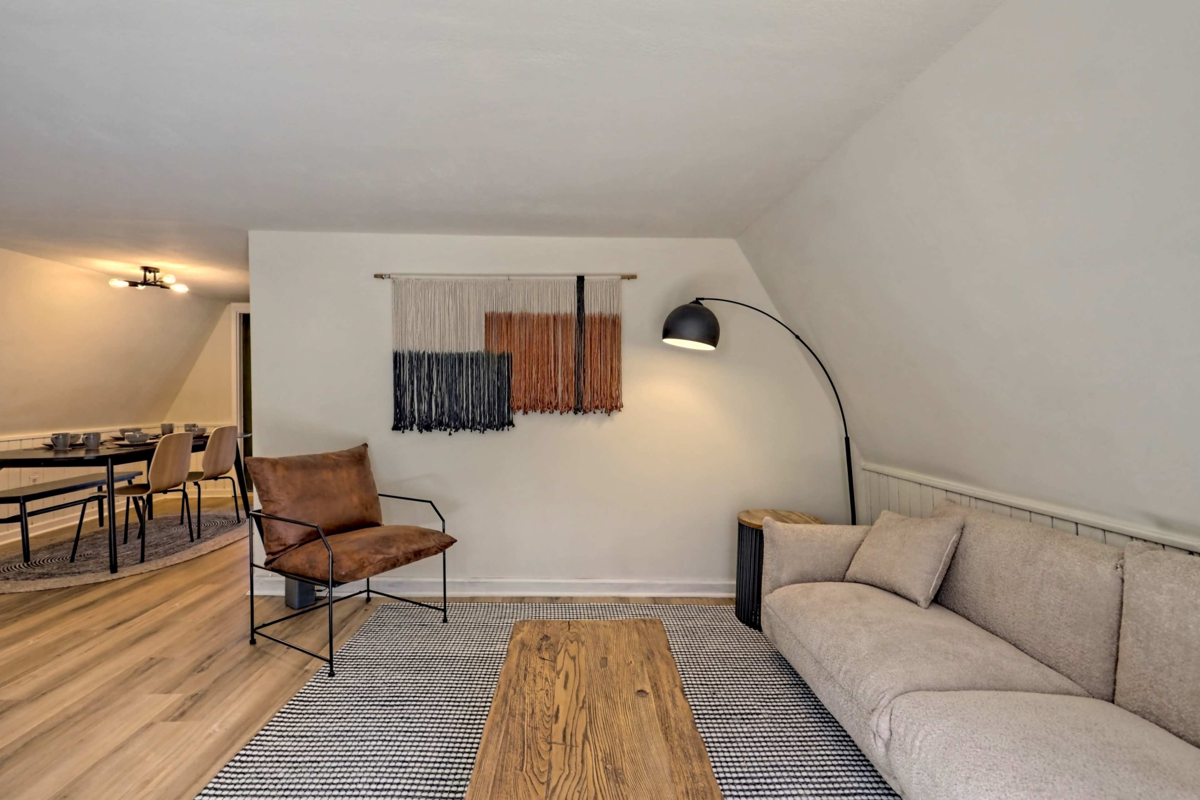 A modern living room featuring a light-colored sofa, an armchair, a wooden coffee table, and a wall hanging above the sofa.
