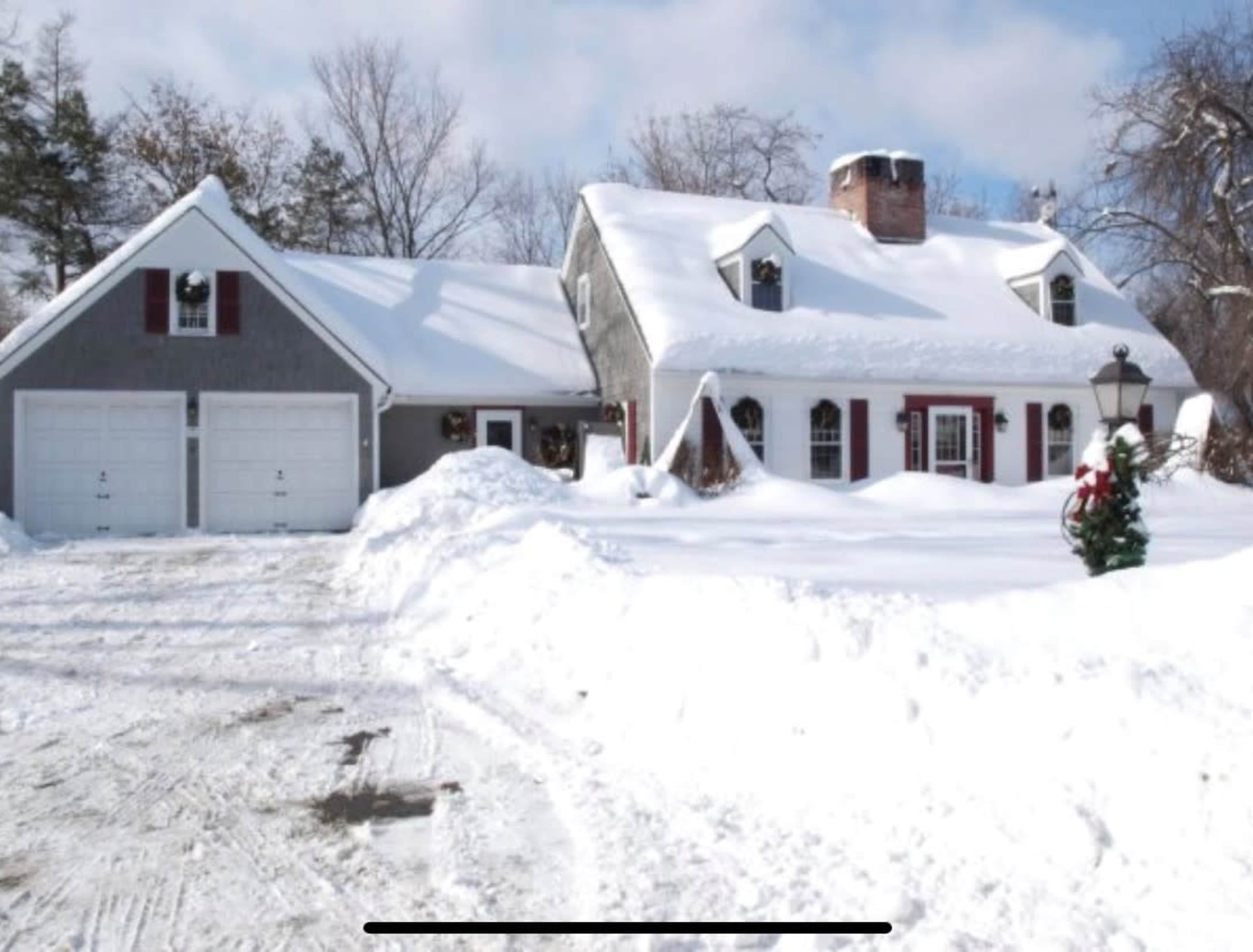 A snow-covered house with a red door and decorative wreaths, flanked by a two-car garage, is surrounded by mounds of snow.