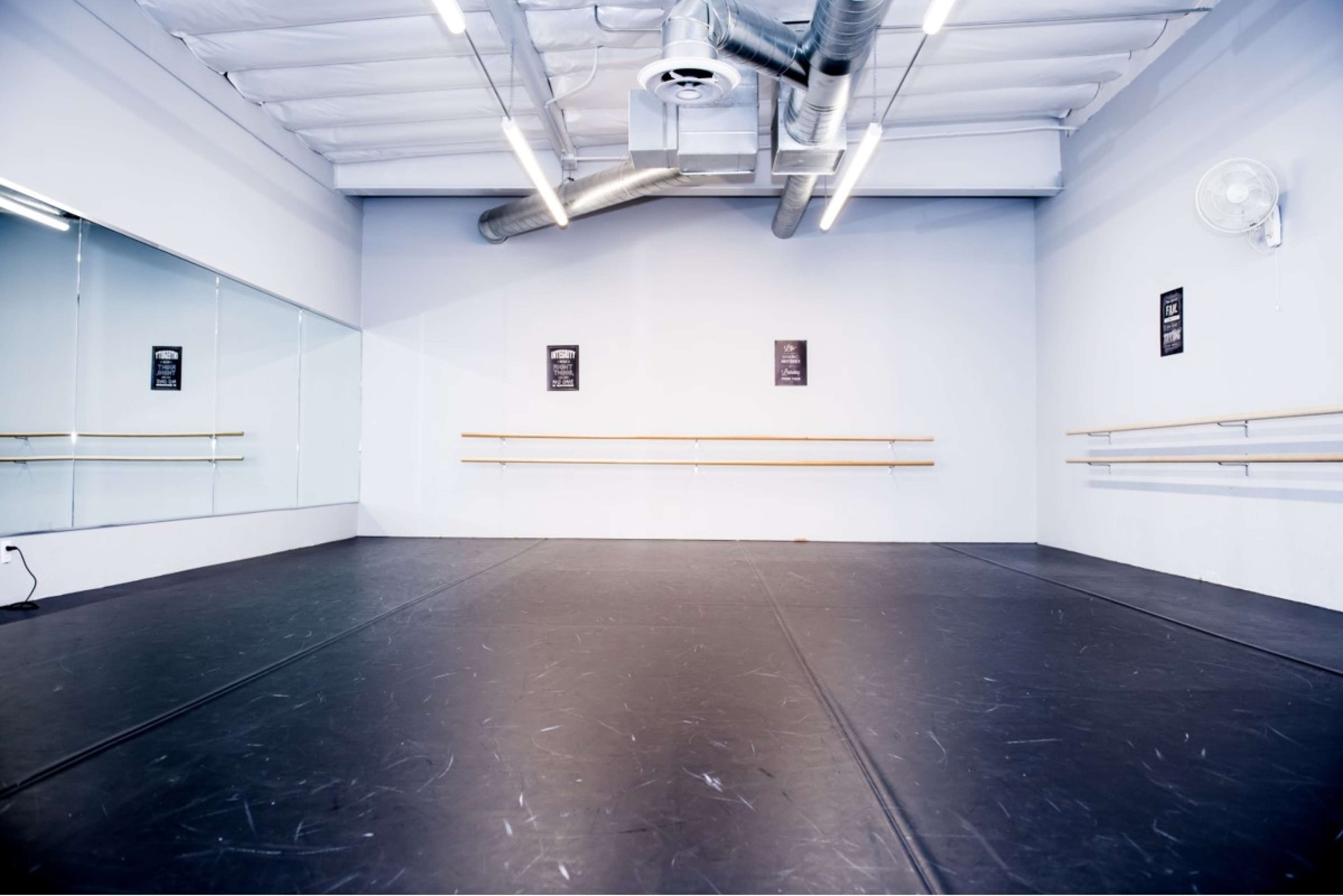 The image shows a spacious dance studio with a wooden ballet bar, mirrors on one wall, and a black rubber floor.