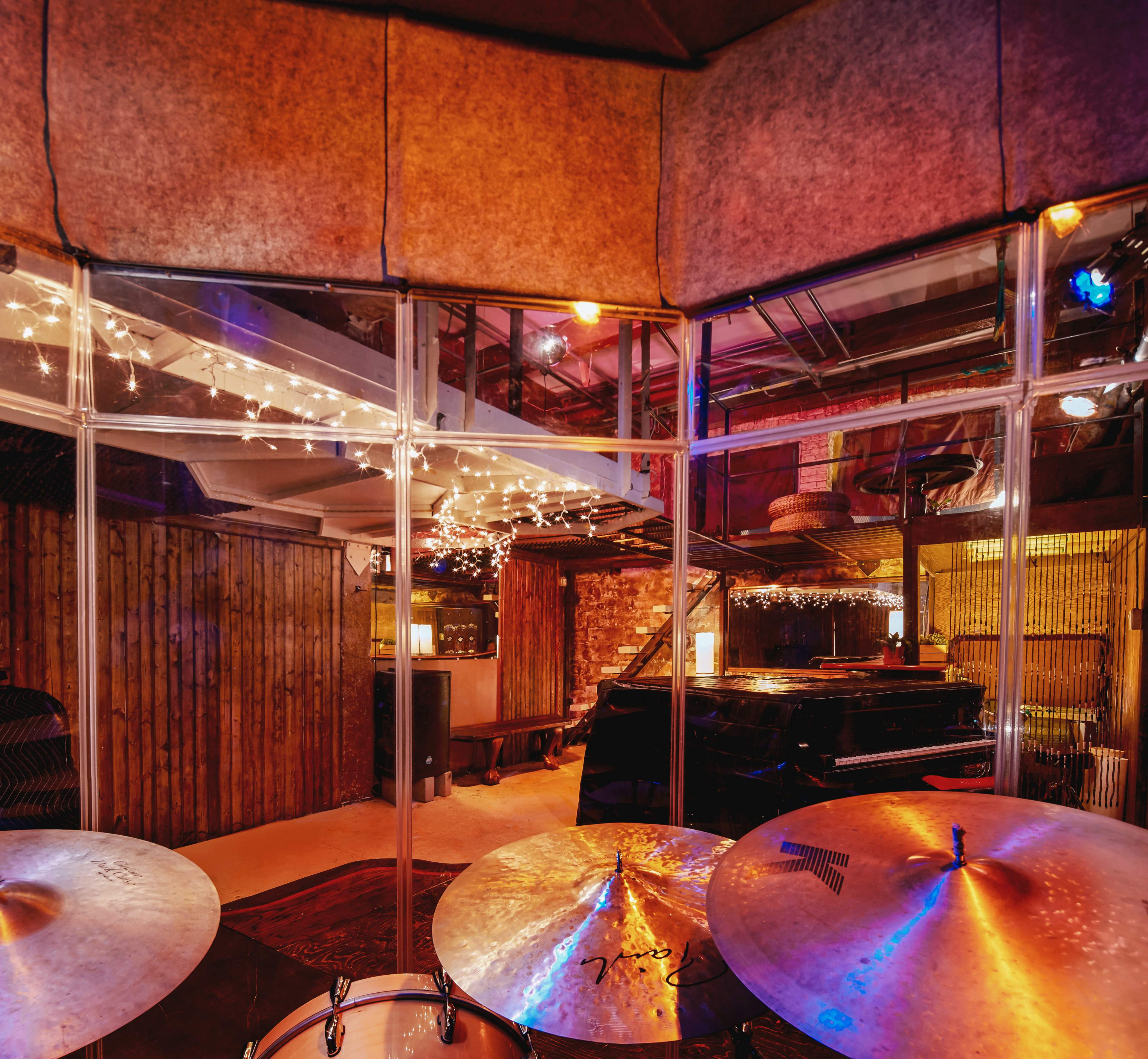A view of a music studio featuring a drum set in the foreground, with a grand piano and string lights visible in the background.