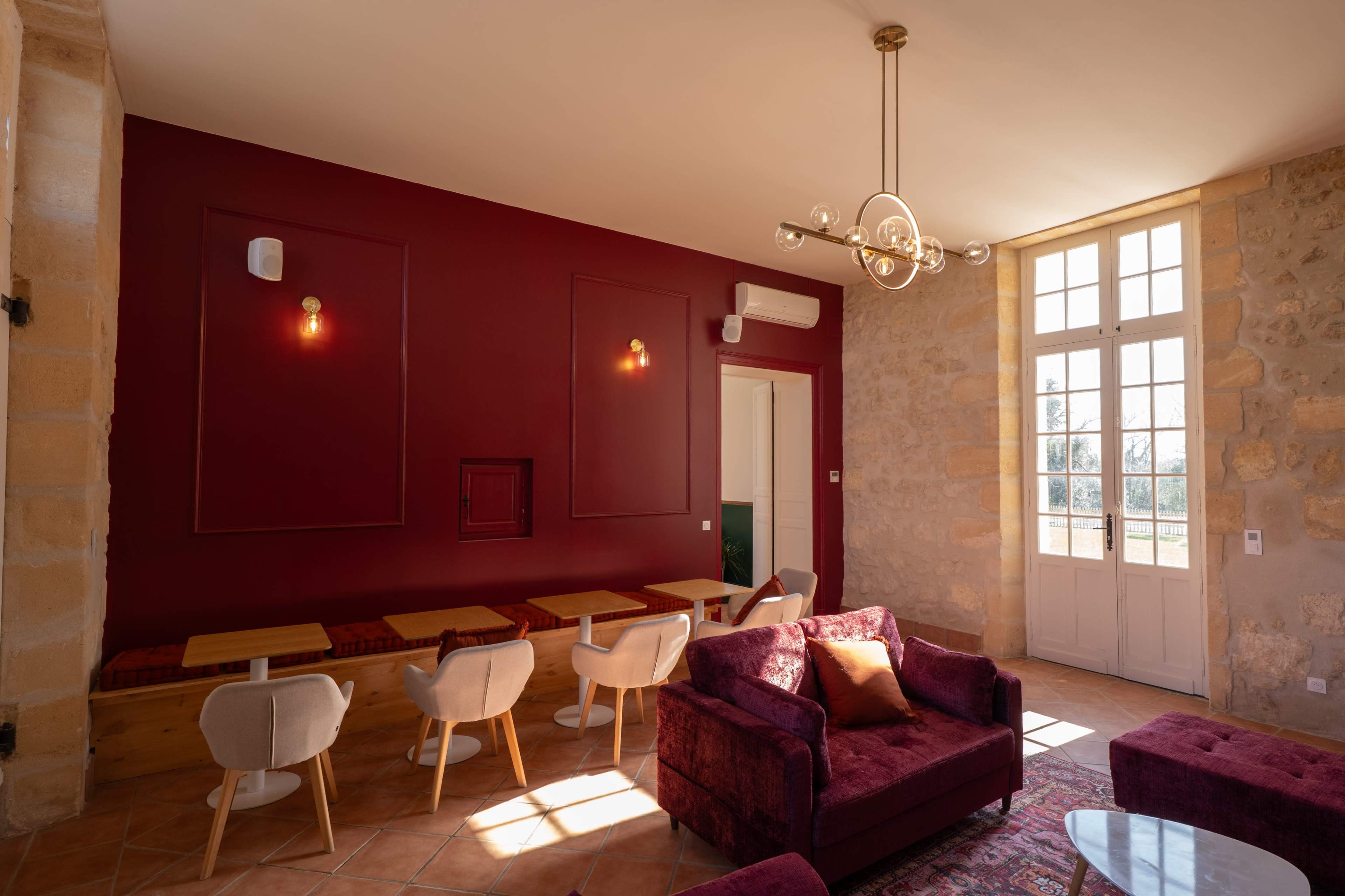 The image shows a cozy interior of a room with stone walls, a dark red accent wall, and a seating area featuring a plush sofa and several small tables.