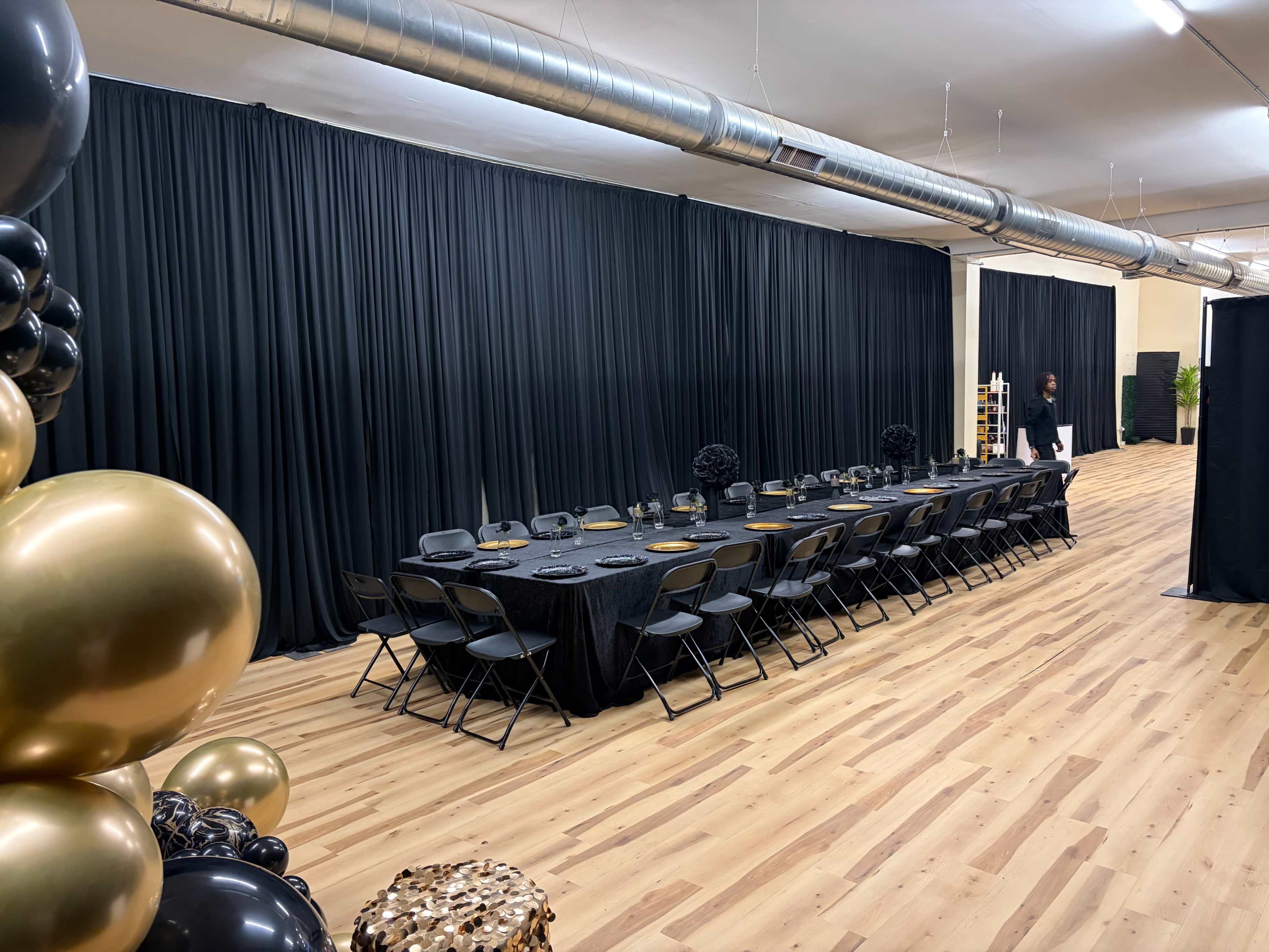 A long, black table with chairs is set up in a spacious room featuring black curtains, wooden flooring, and decorative balloons.