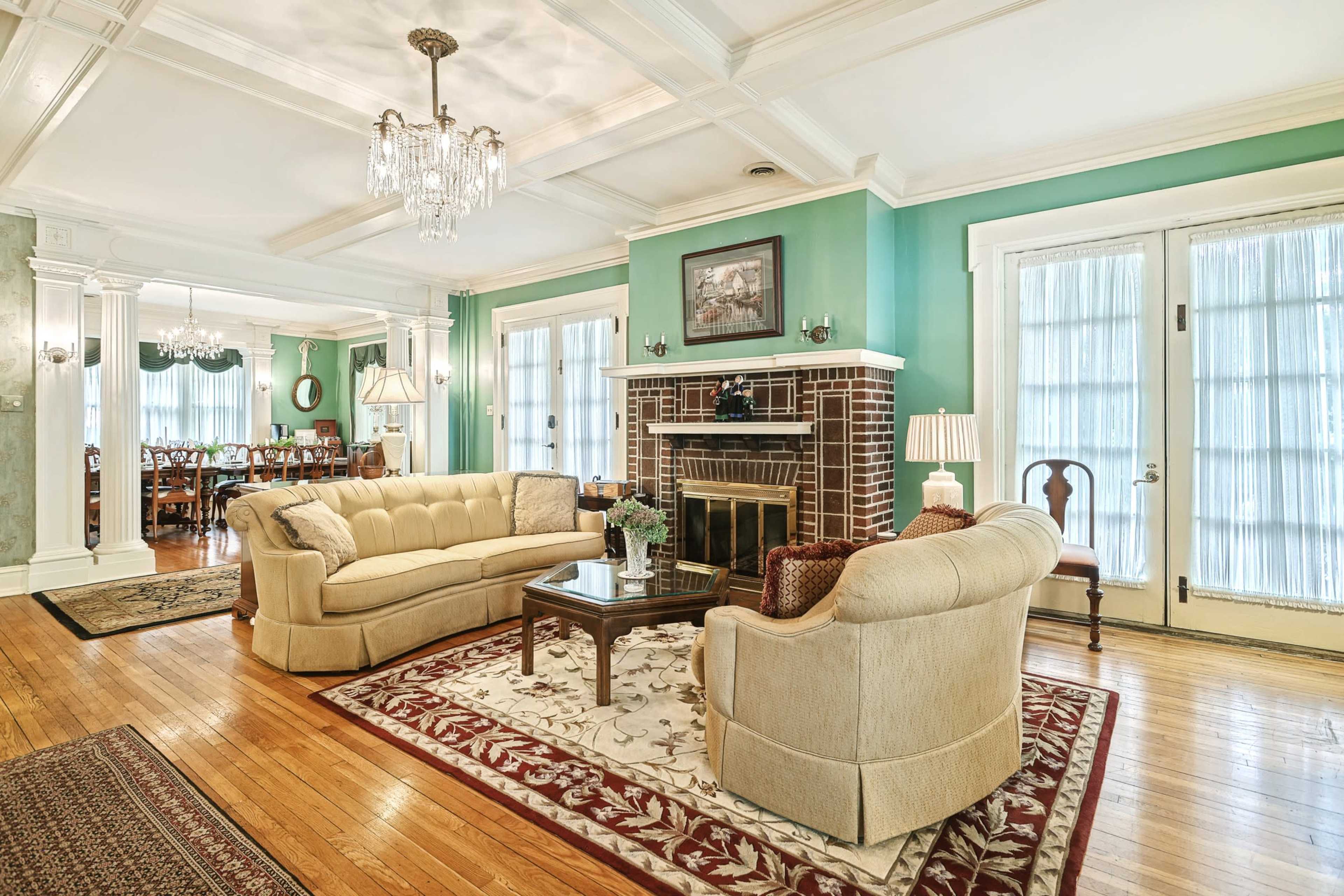 A bright living room with a beige couch, a coffee table, and a brick fireplace, featuring large windows and a decorative rug.