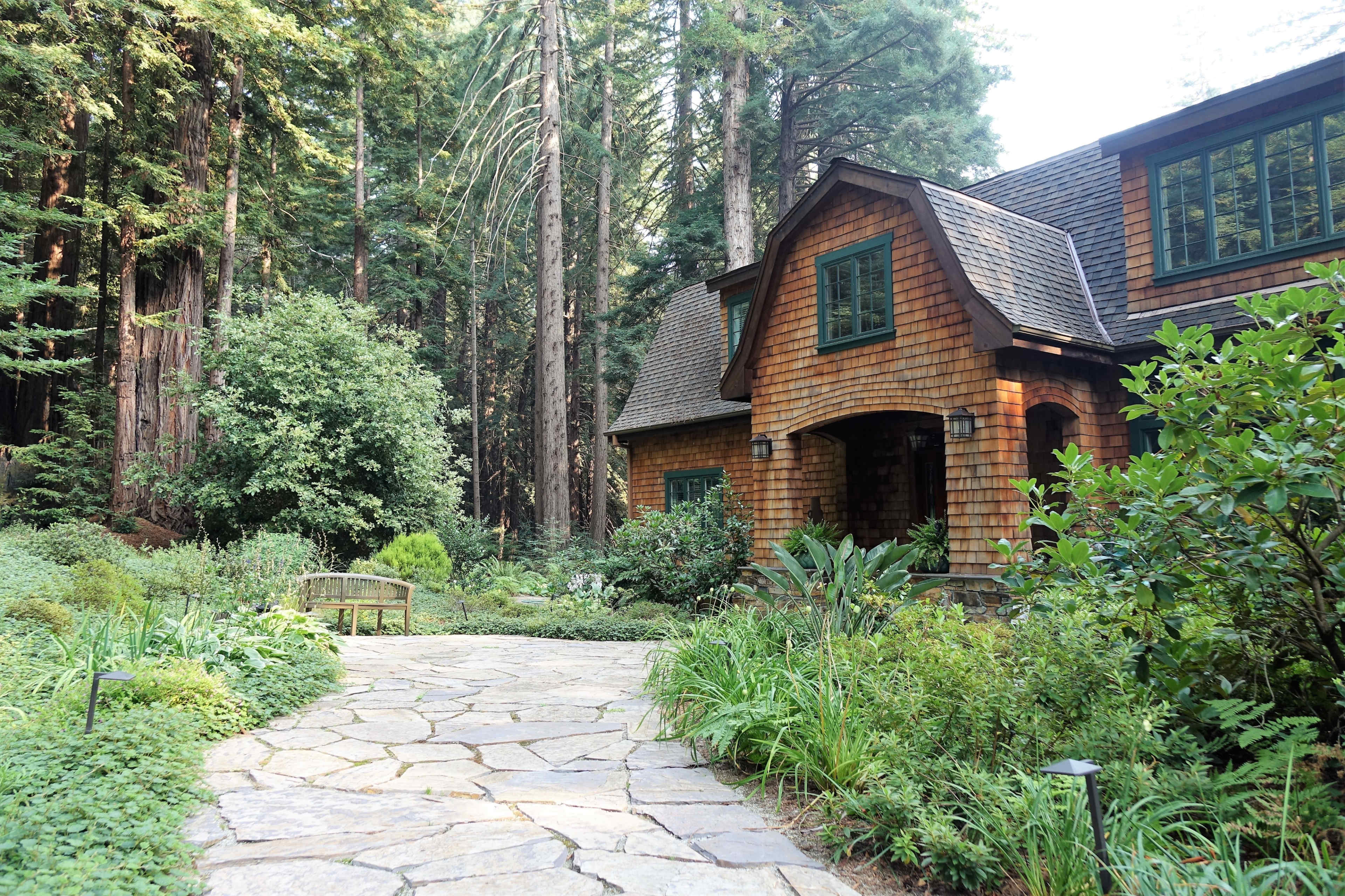 A stone pathway leads to a wooden house surrounded by lush greenery and tall trees.