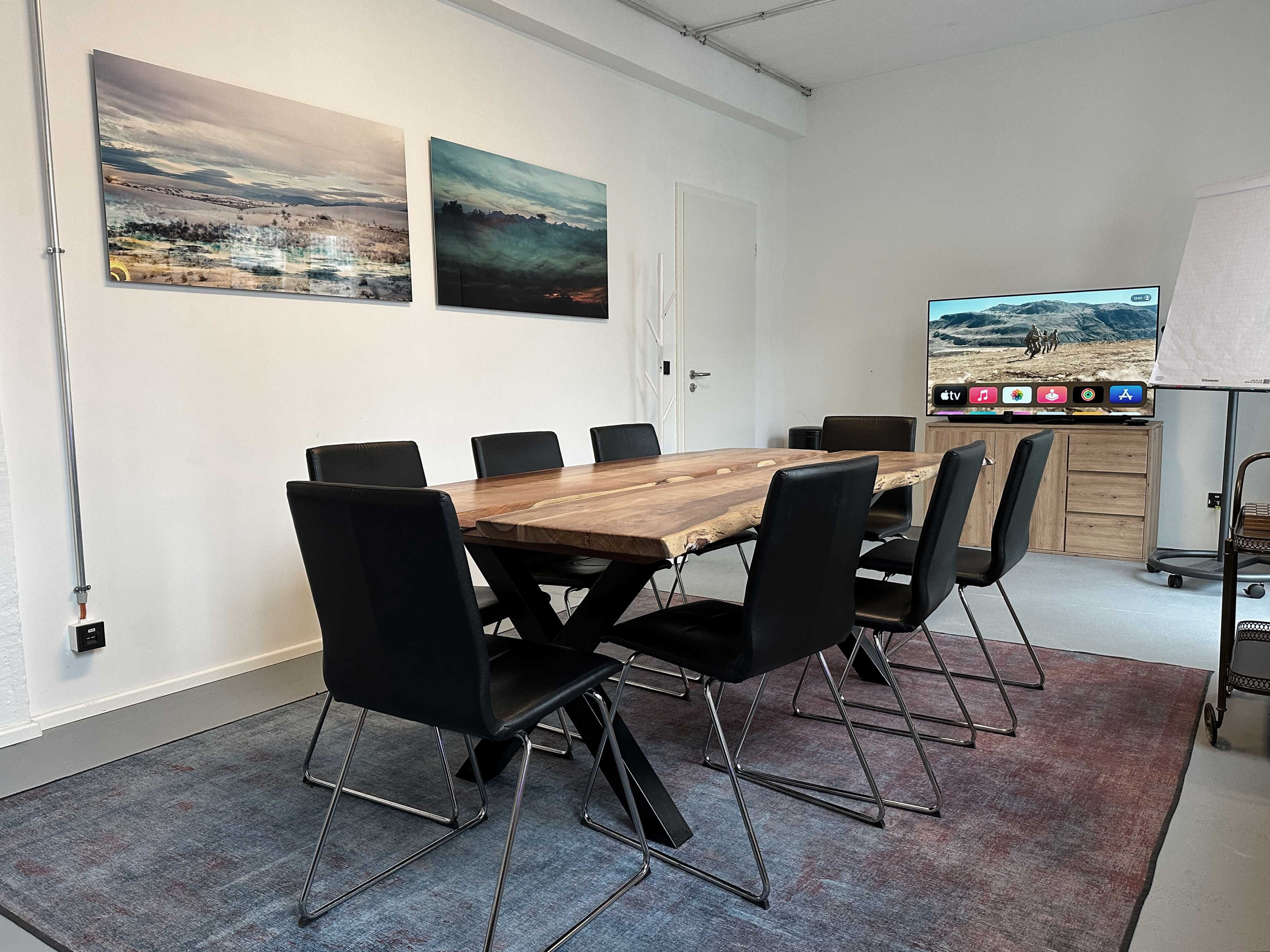 A modern conference room features a wooden table surrounded by black chairs, with two landscape paintings on the wall and a television on a stand.