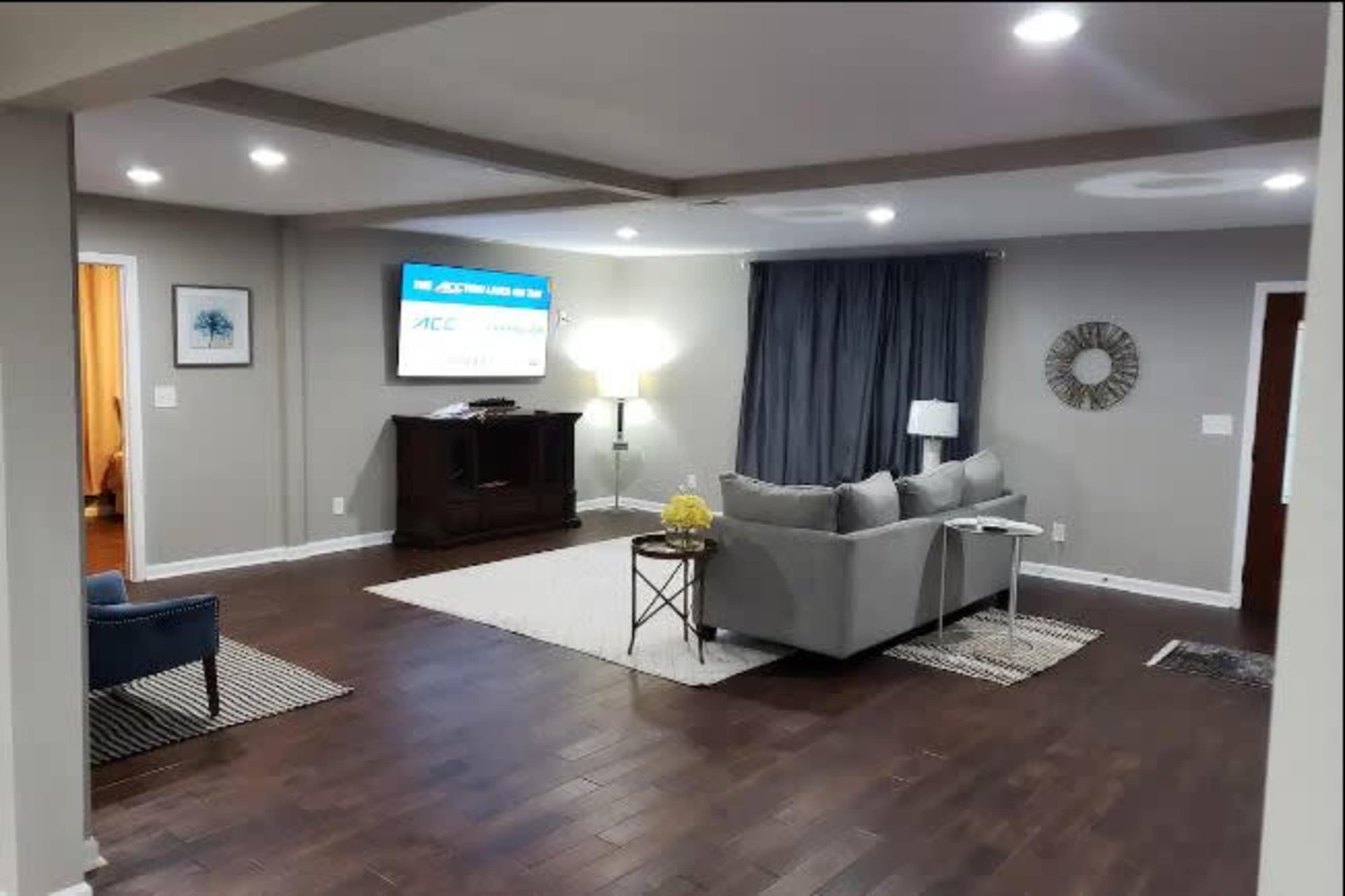 The living room features a gray sofa, a coffee table, a dark wooden cabinet, and a large screen mounted on the wall, with warm lighting and a mix of hardwood and rug flooring.