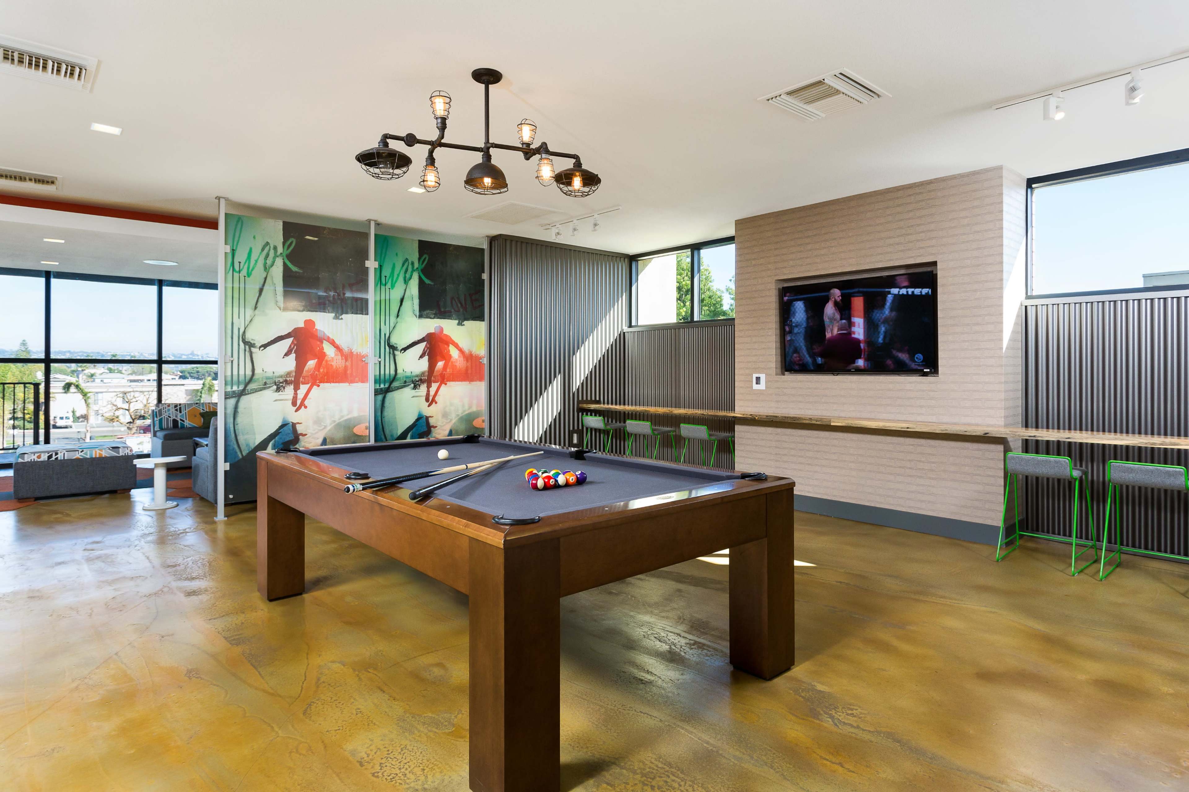 The image shows a modern game room featuring a pool table, a large television, and a seating area with floor-to-ceiling windows offering a city view.