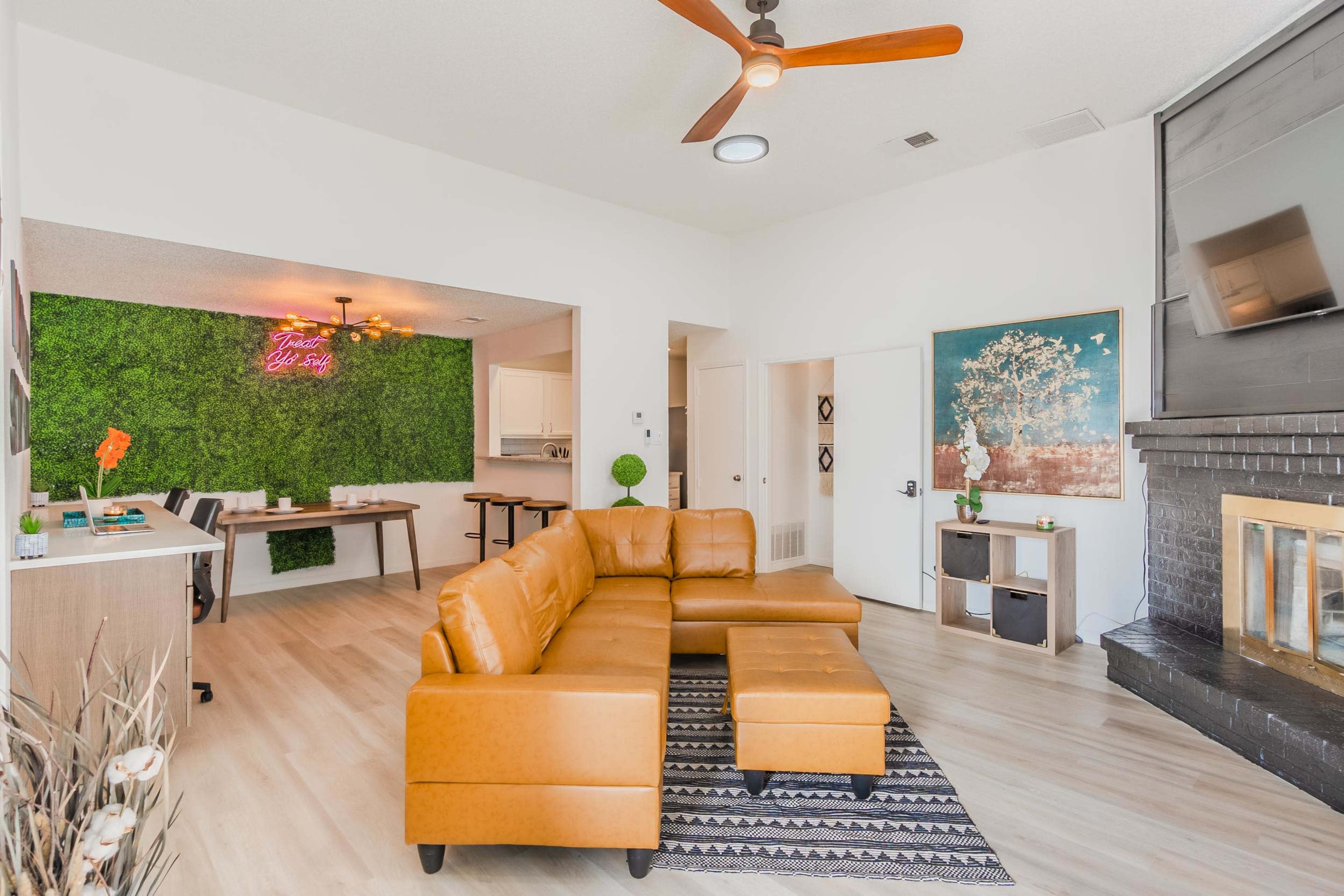 The image shows a modern living room with a brown sectional sofa, a fireplace, a dining area with a table, and a green wall feature.