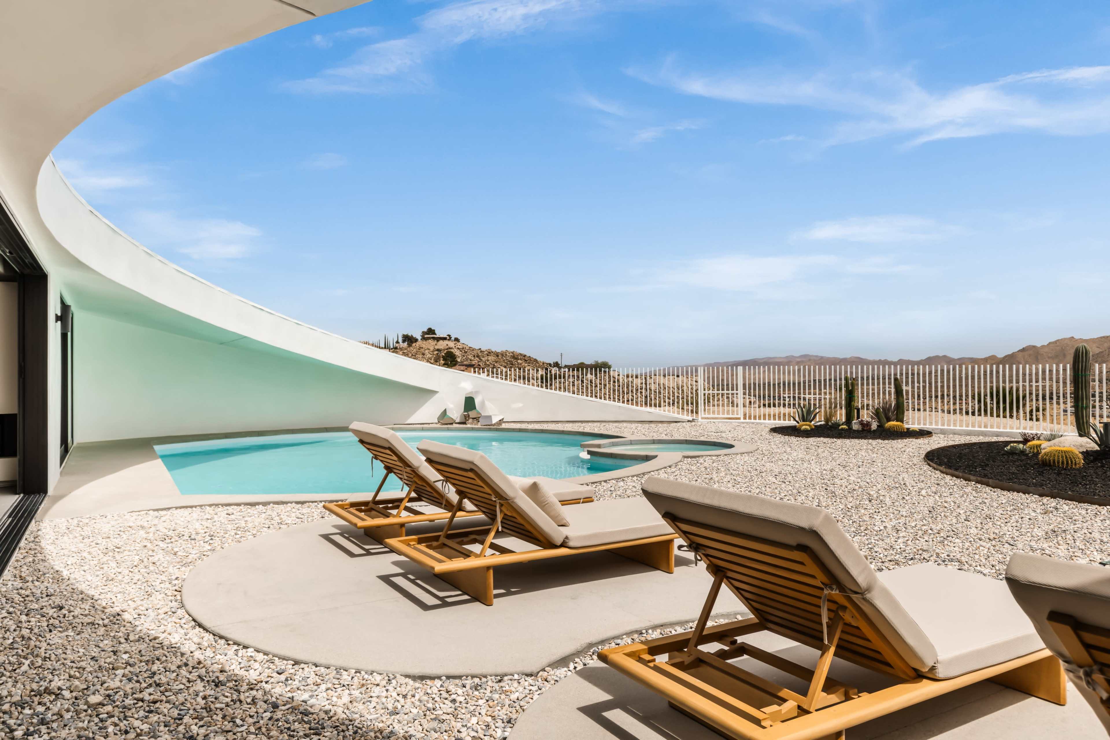 The image shows a modern outdoor space featuring a swimming pool, sun loungers, and a desert landscape in the background.