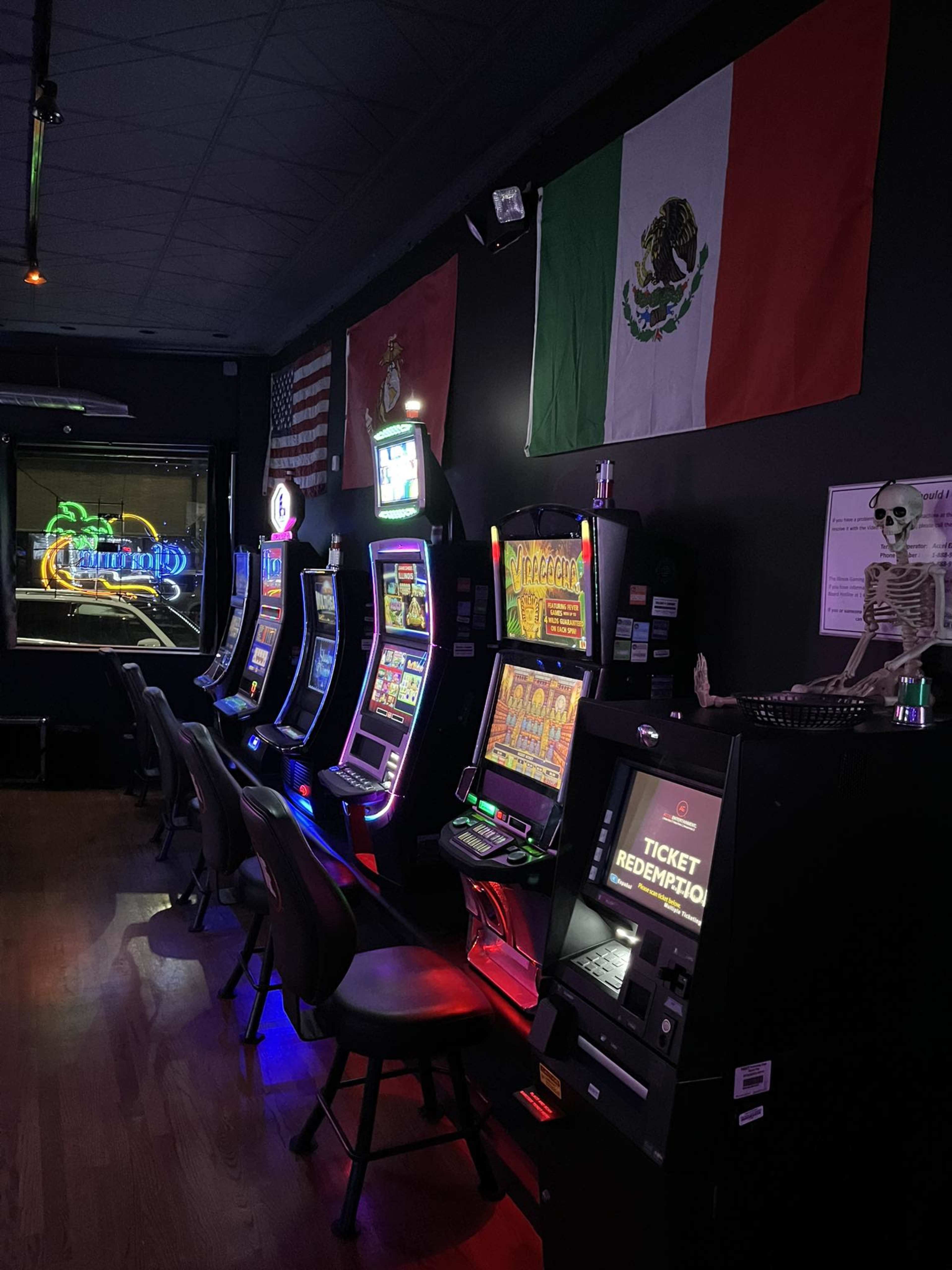 A row of slot machines in a dimly lit room, with Mexican and American flags hanging on the wall.