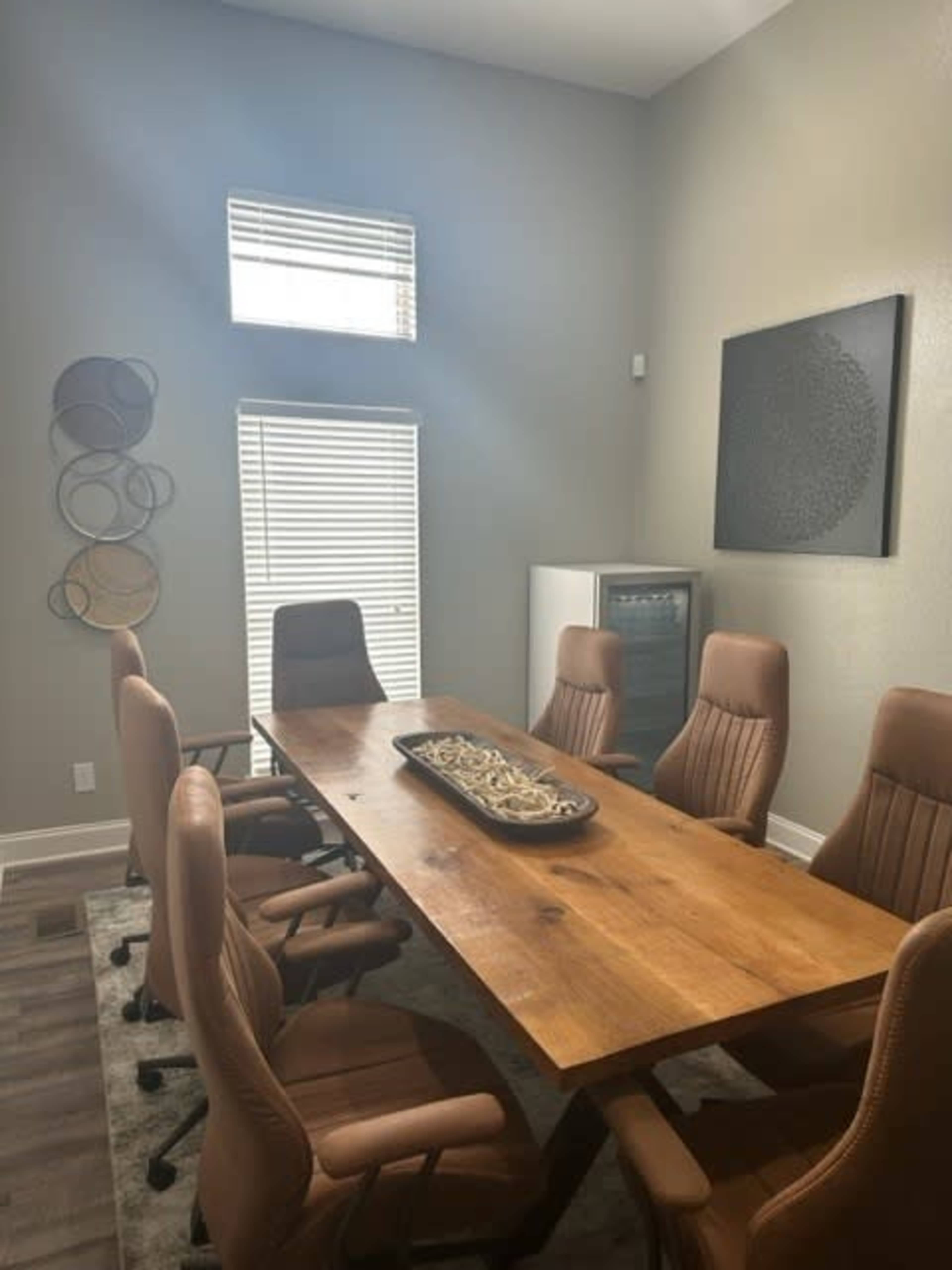 A conference room features a large wooden table surrounded by six brown leather chairs, with a decorative platter in the center and a large window allowing natural light.