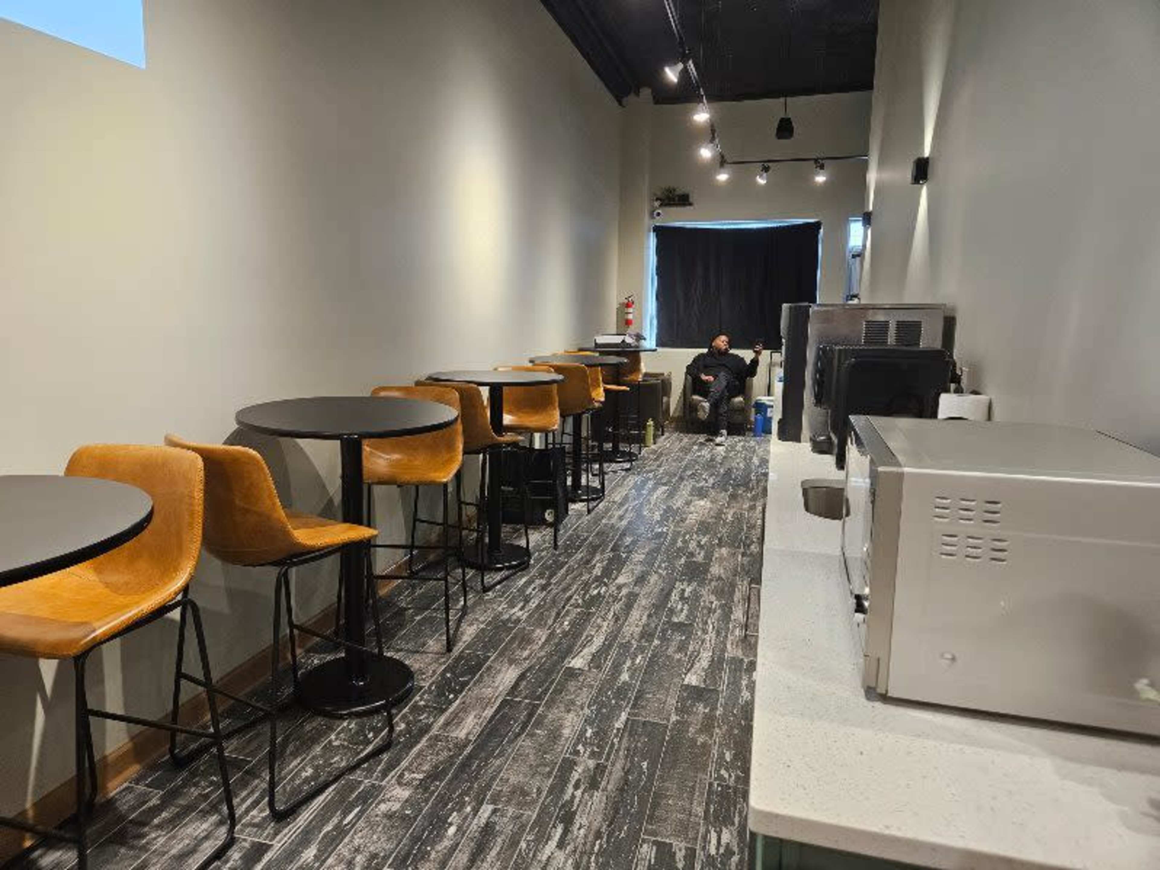 The image shows a narrow café interior with several round tables and chairs along one side, and a counter area with kitchen appliances on the other side, while a lone customer is seated at the far end.
