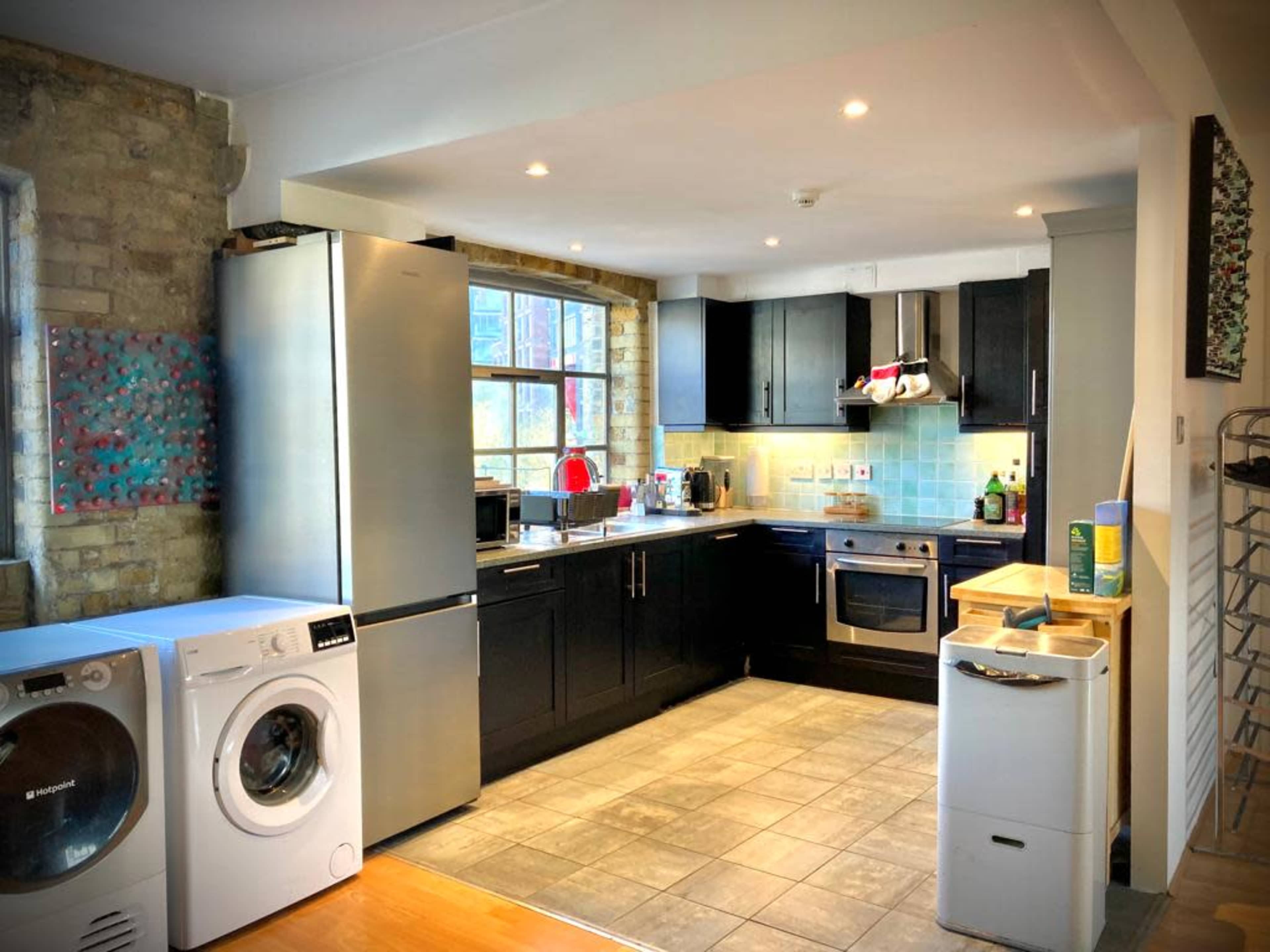 A modern kitchen with stainless steel appliances, dark cabinetry, and tiled flooring, featuring a washer and a bright window.