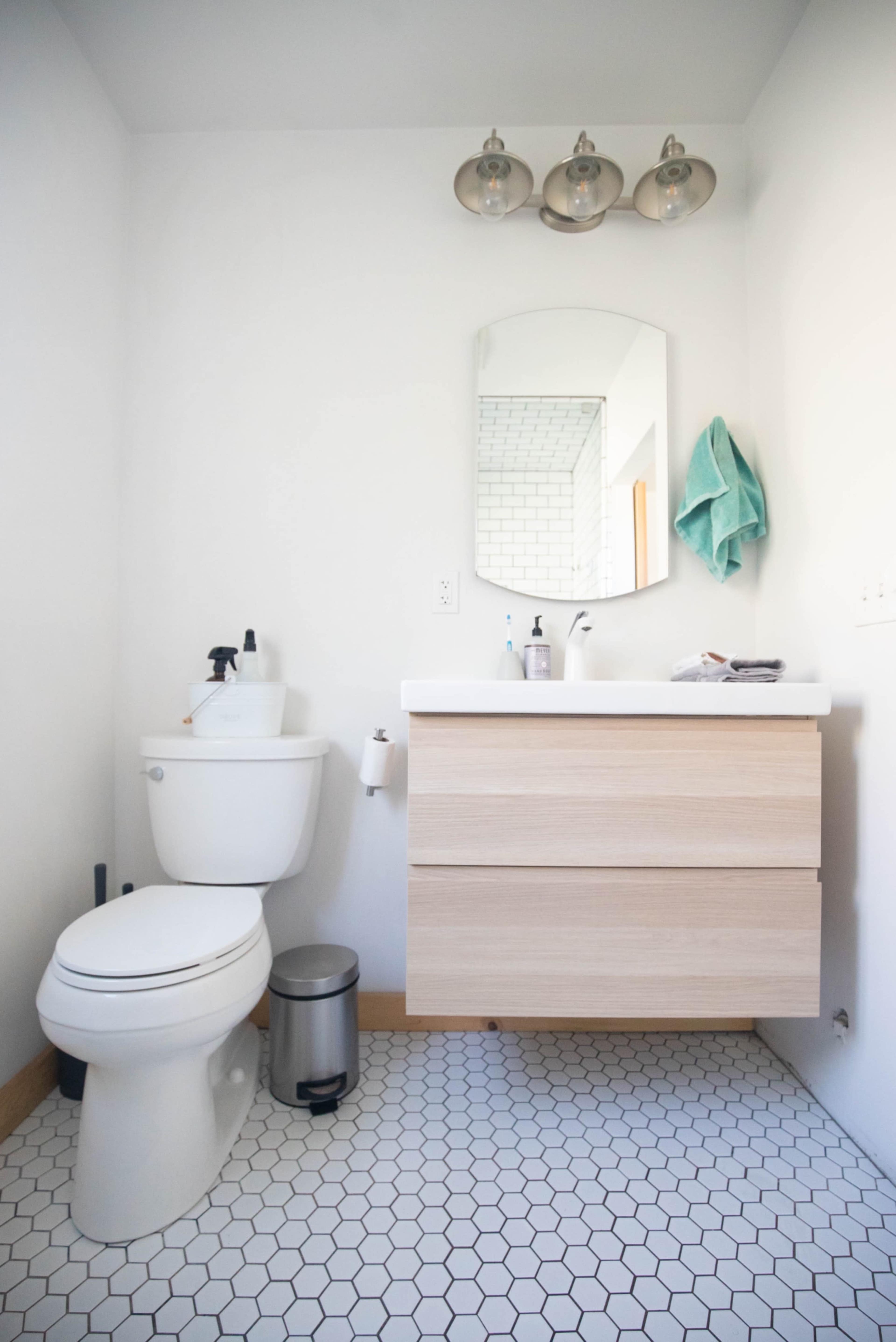 A modern bathroom features a white wall, a mirror above a minimalist wooden vanity with drawers, a toilet, and a small trash can on a hexagonal tiled floor.