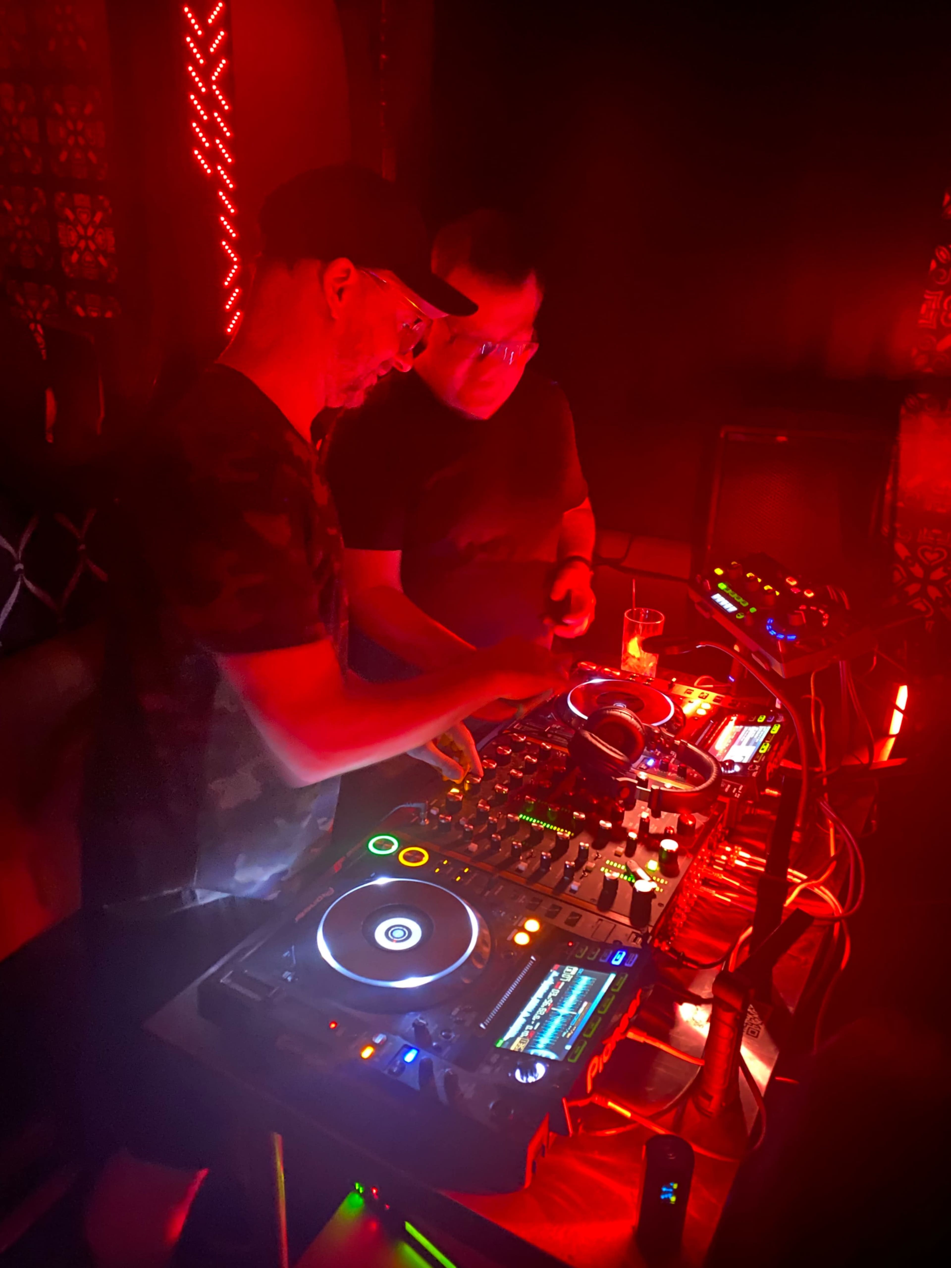 Two DJs are mixing music at a club, surrounded by colorful red lighting.