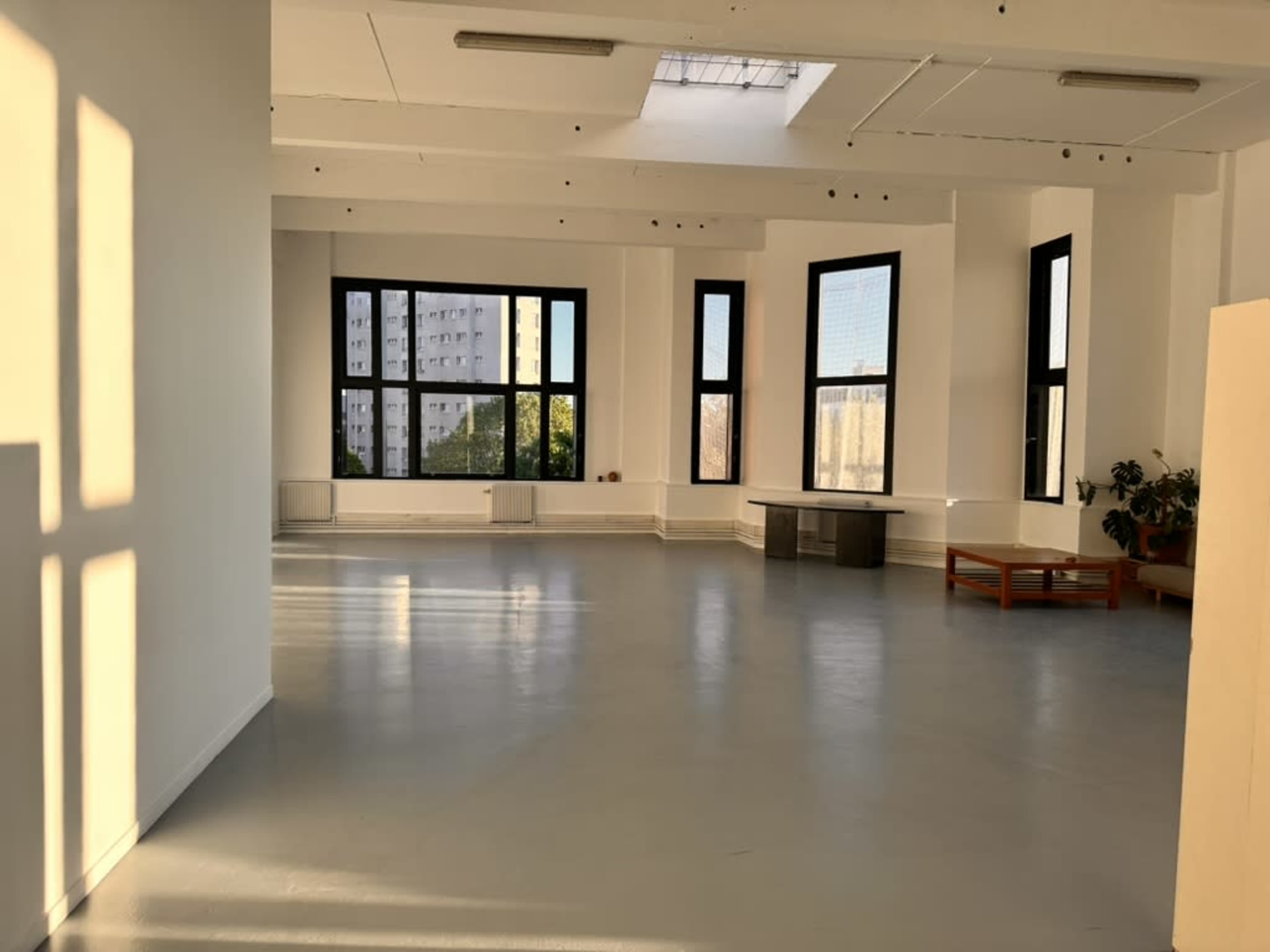 The image shows a spacious, sunlit room with large windows, a light-colored floor, and minimal furniture.