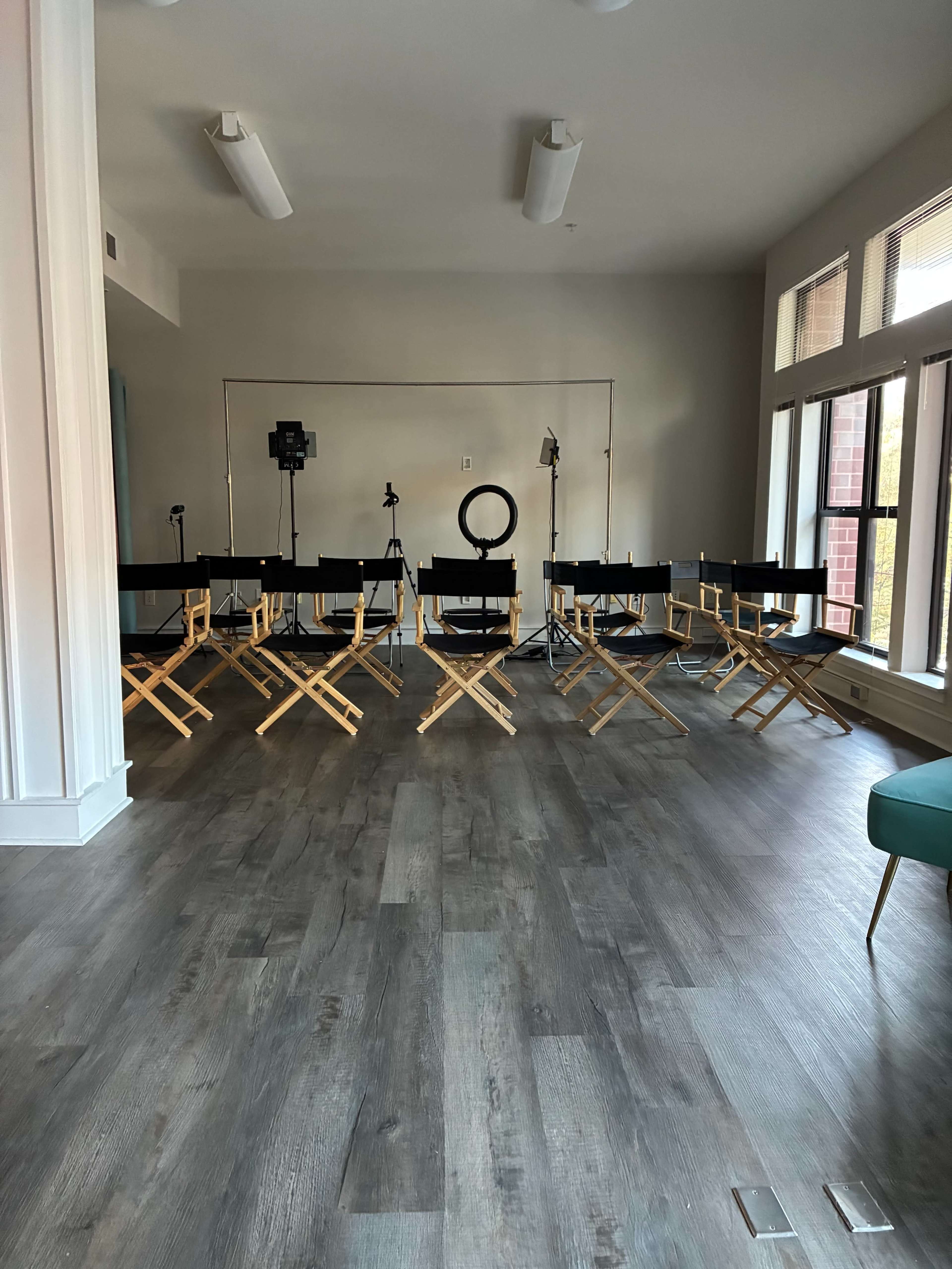 The image shows a spacious room with wooden flooring, featuring several director's chairs arranged in a semi-circle around a central lighting setup.