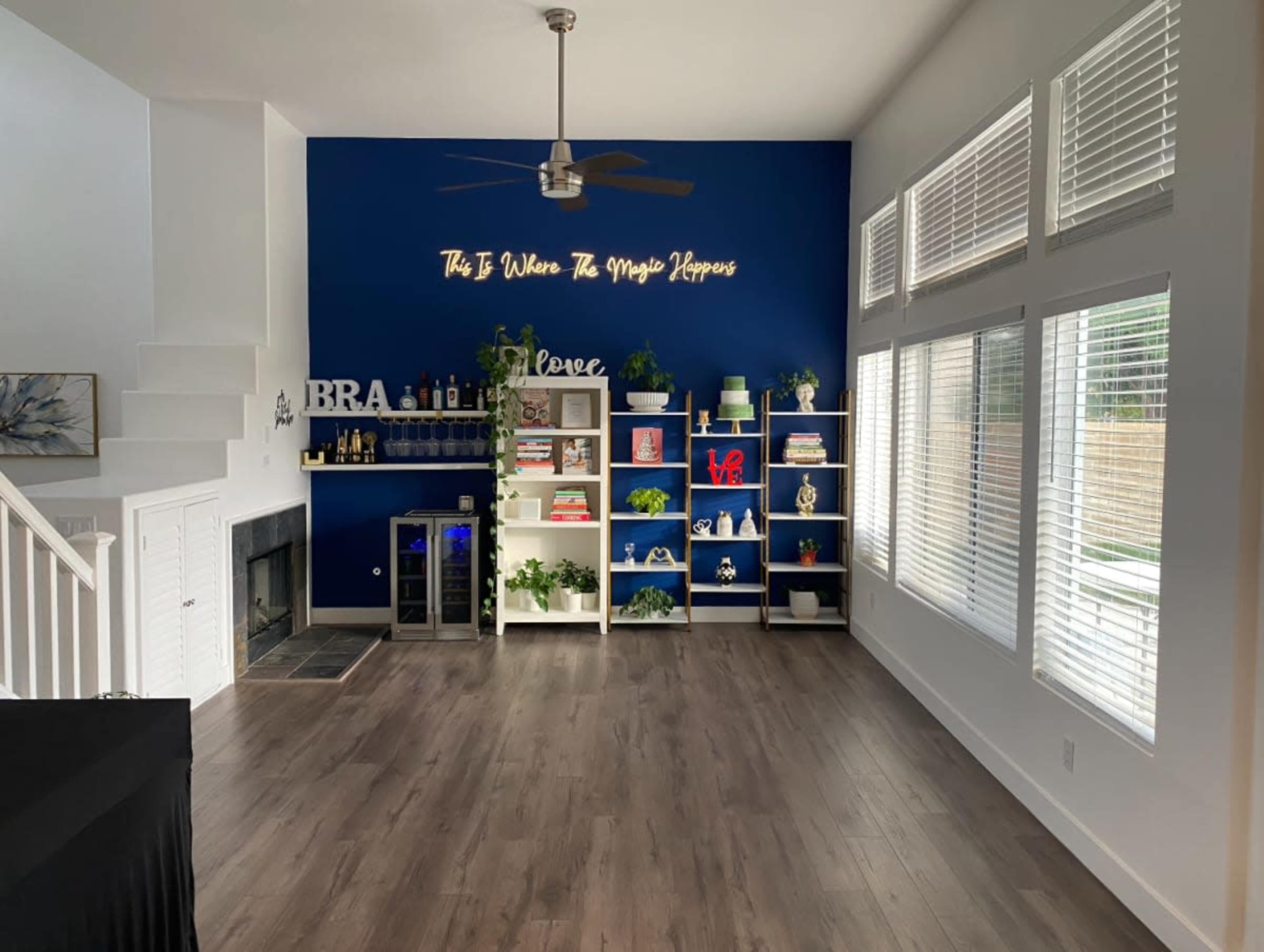 The image shows a modern living space with blue and white walls, featuring a fireplace, shelves filled with decorative items and plants, and a sign that reads "This Is Where The Magic Happens."