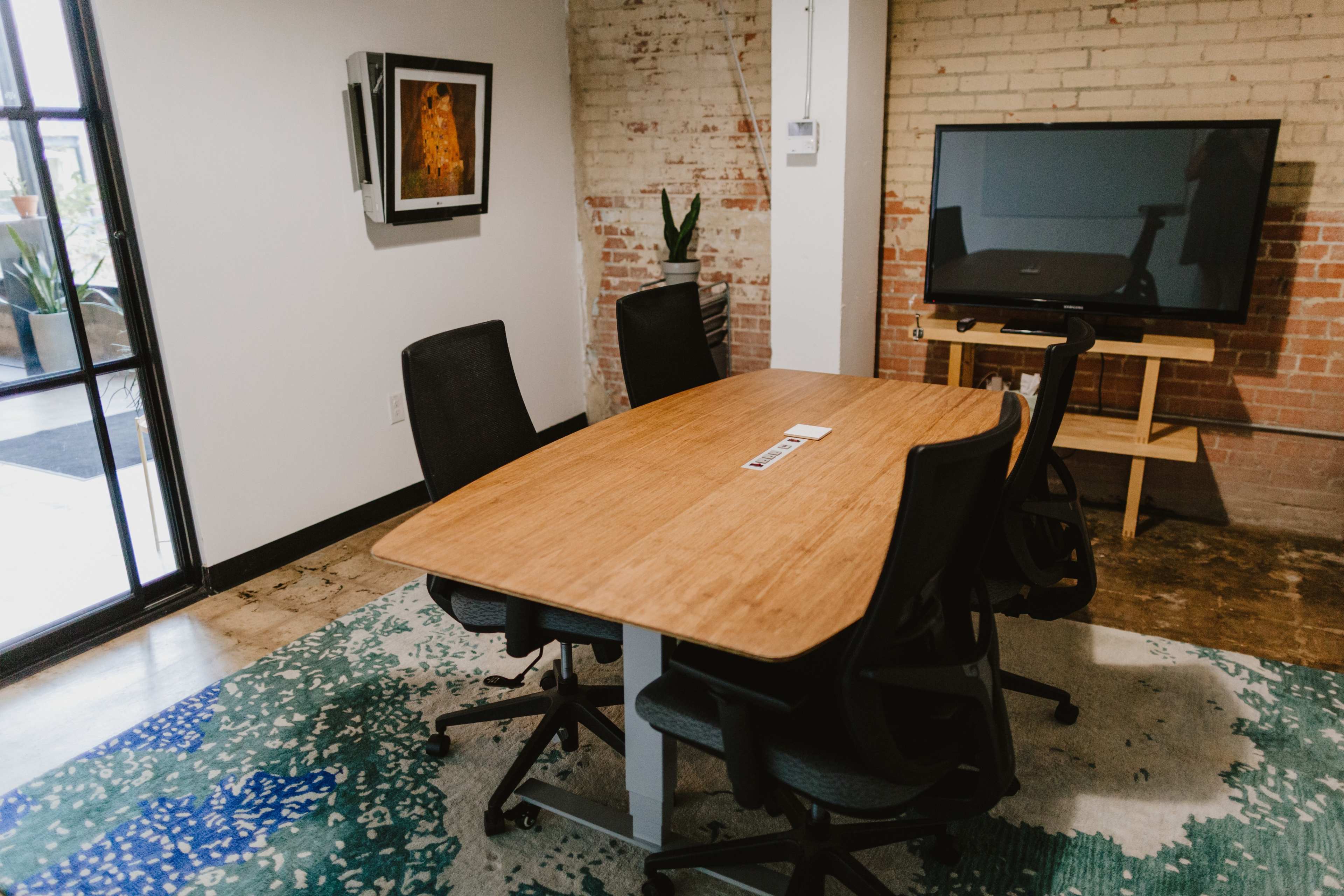 A modern conference room features a wooden table surrounded by five black chairs, a television mounted on the wall, and brick accents.
