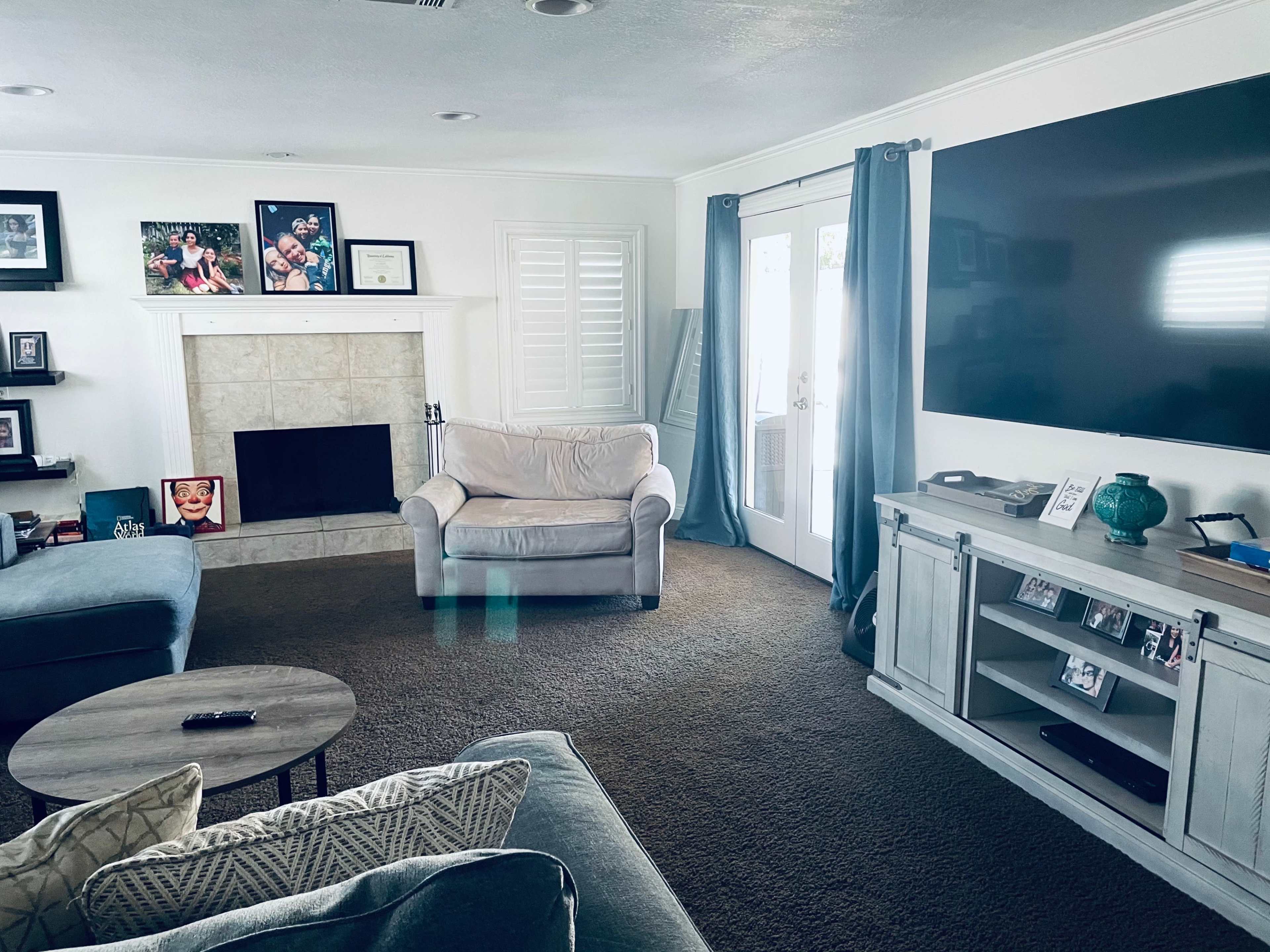 The living room features a beige armchair, a wooden coffee table, a fireplace with framed photos, and a large television mounted on the wall.