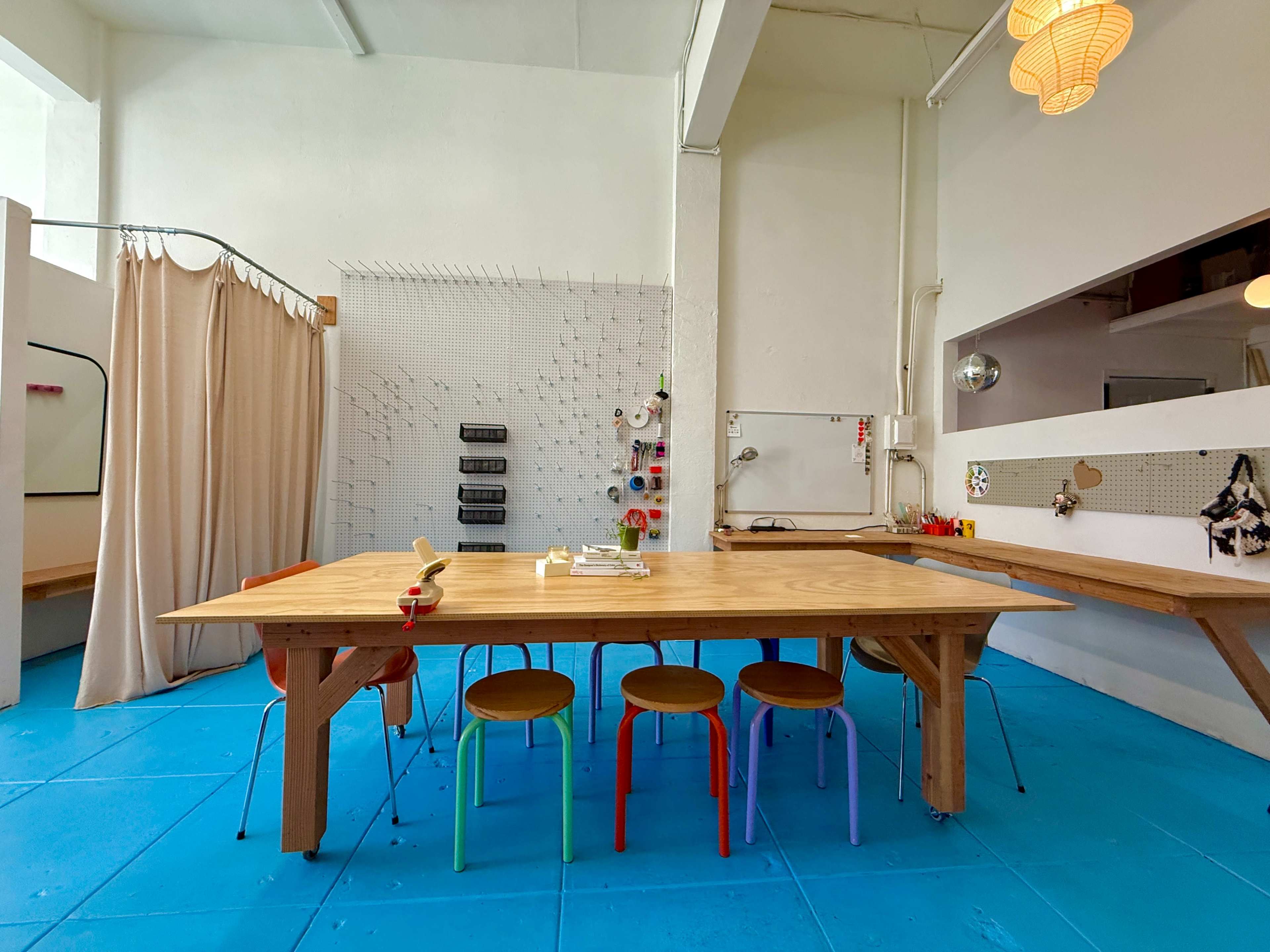 A spacious, brightly lit room features a large wooden table surrounded by colorful chairs, with a backdrop of organized tools and materials on the walls.