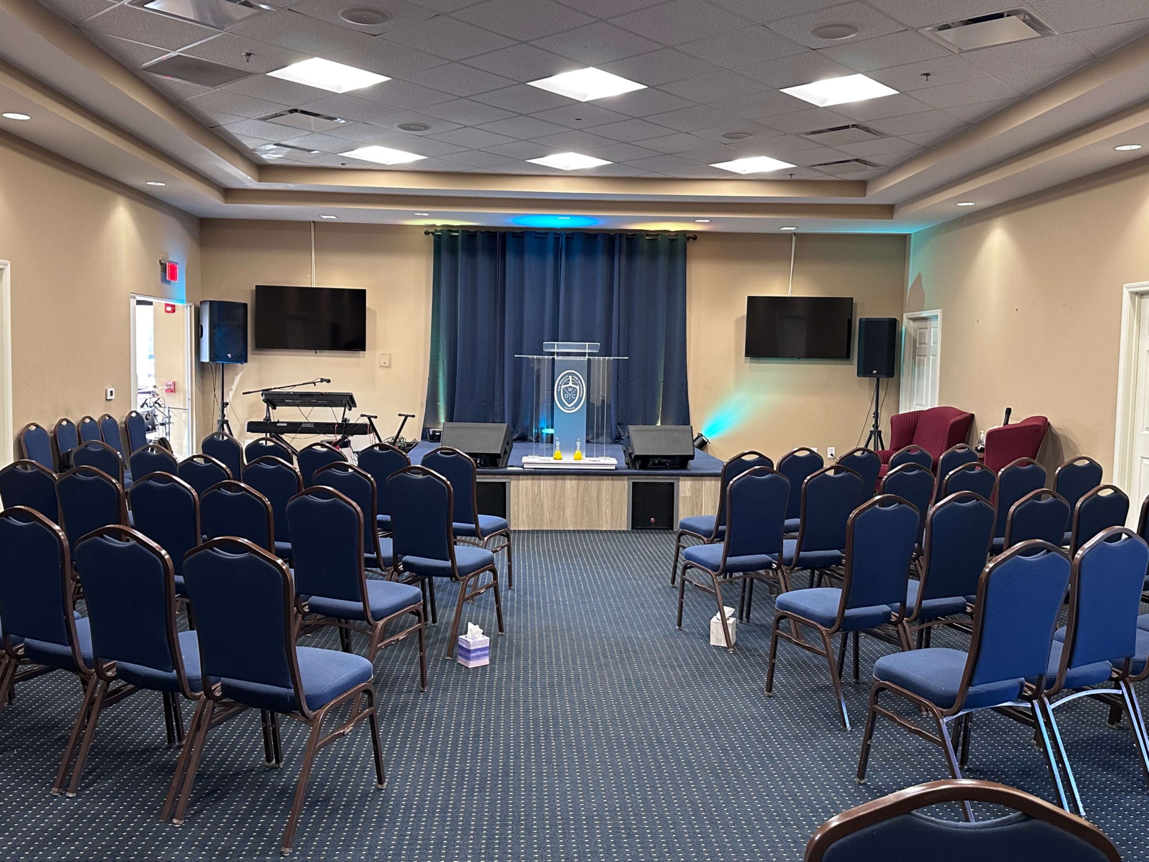 The image shows a spacious church interior with rows of blue chairs facing a stage that has a podium and two television screens.