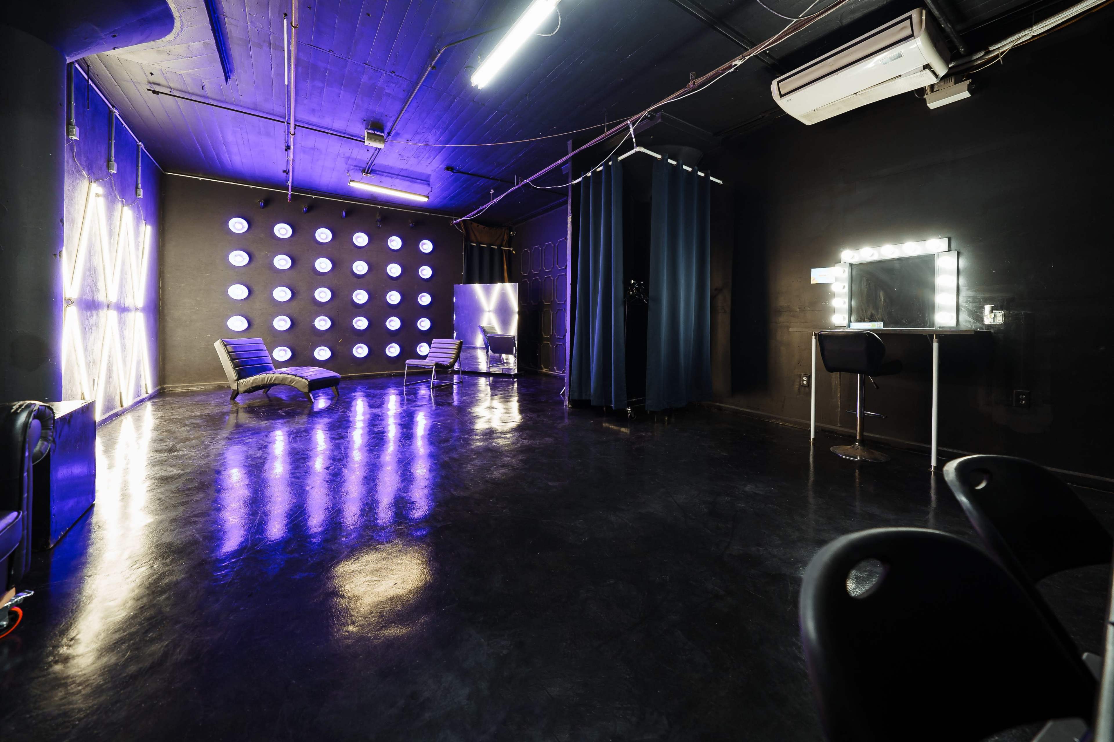 A modern, dimly lit room with black walls, illuminated by purple and white light fixtures, featuring lounge chairs, a makeup station with mirrors, and a changing area.