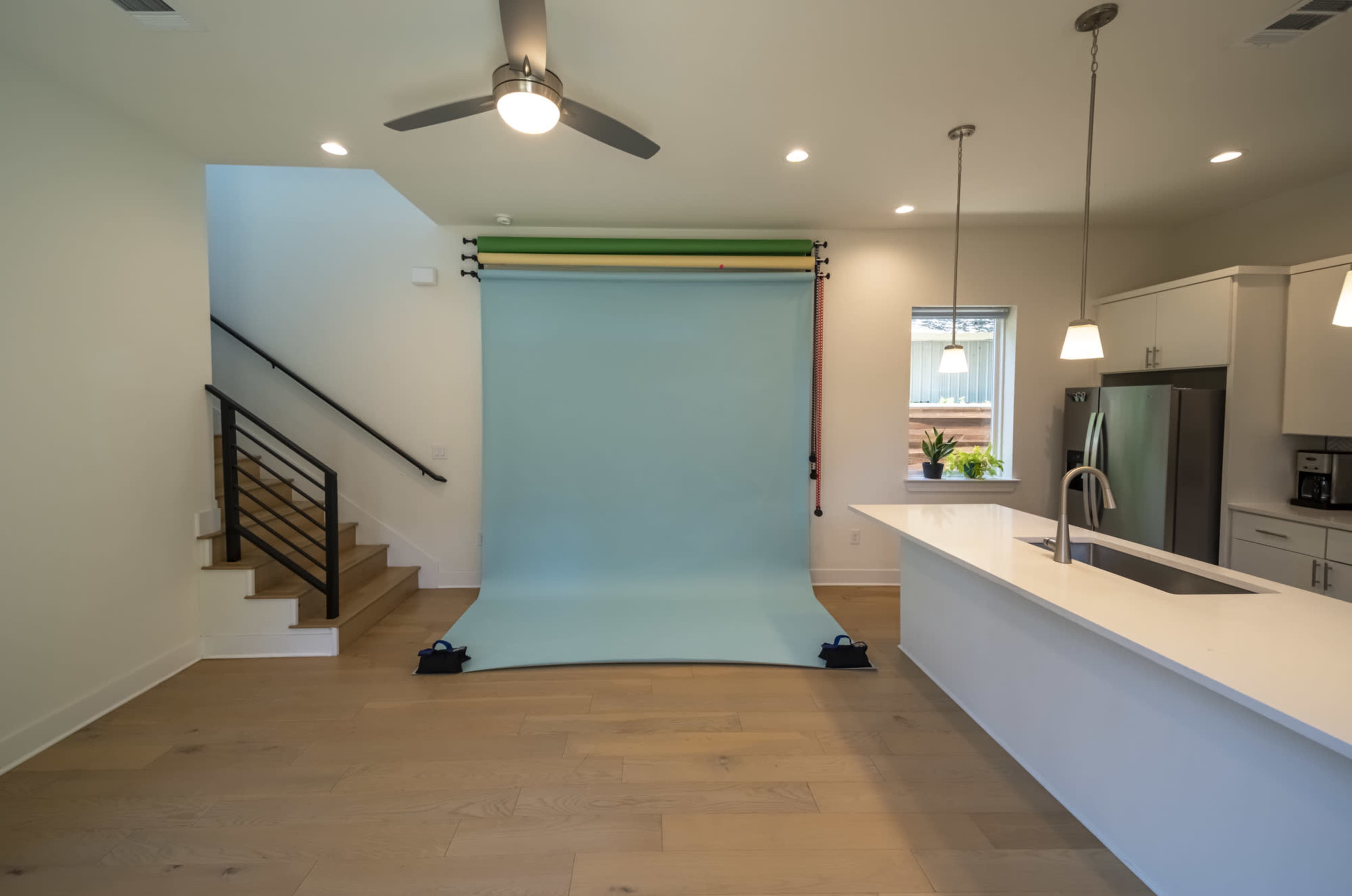 The image shows a modern kitchen and living area featuring a light blue photo backdrop and a staircase in the background.