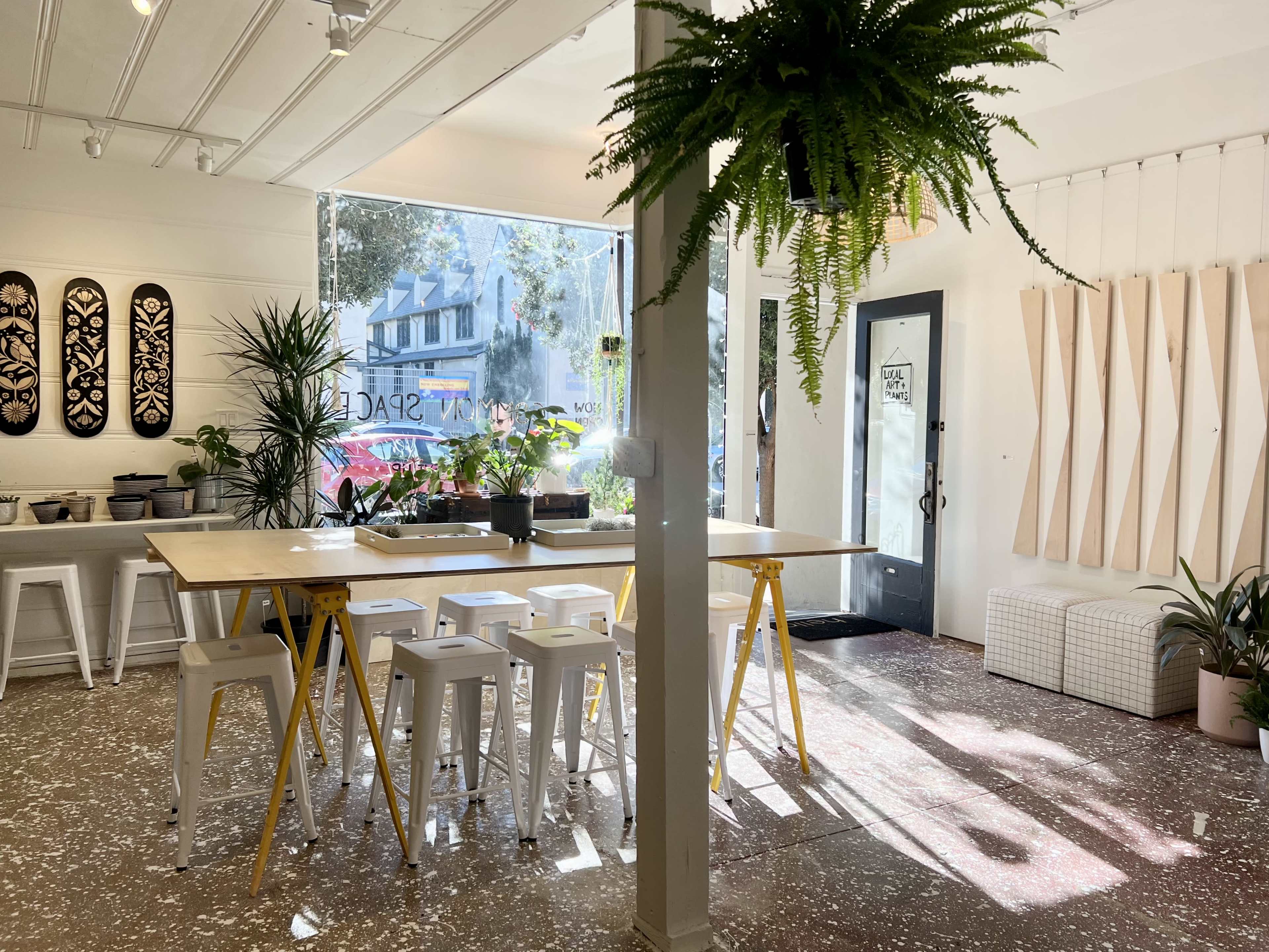 A bright interior of a contemporary space with a long wooden table surrounded by white stools, plants, and large windows allowing natural light.