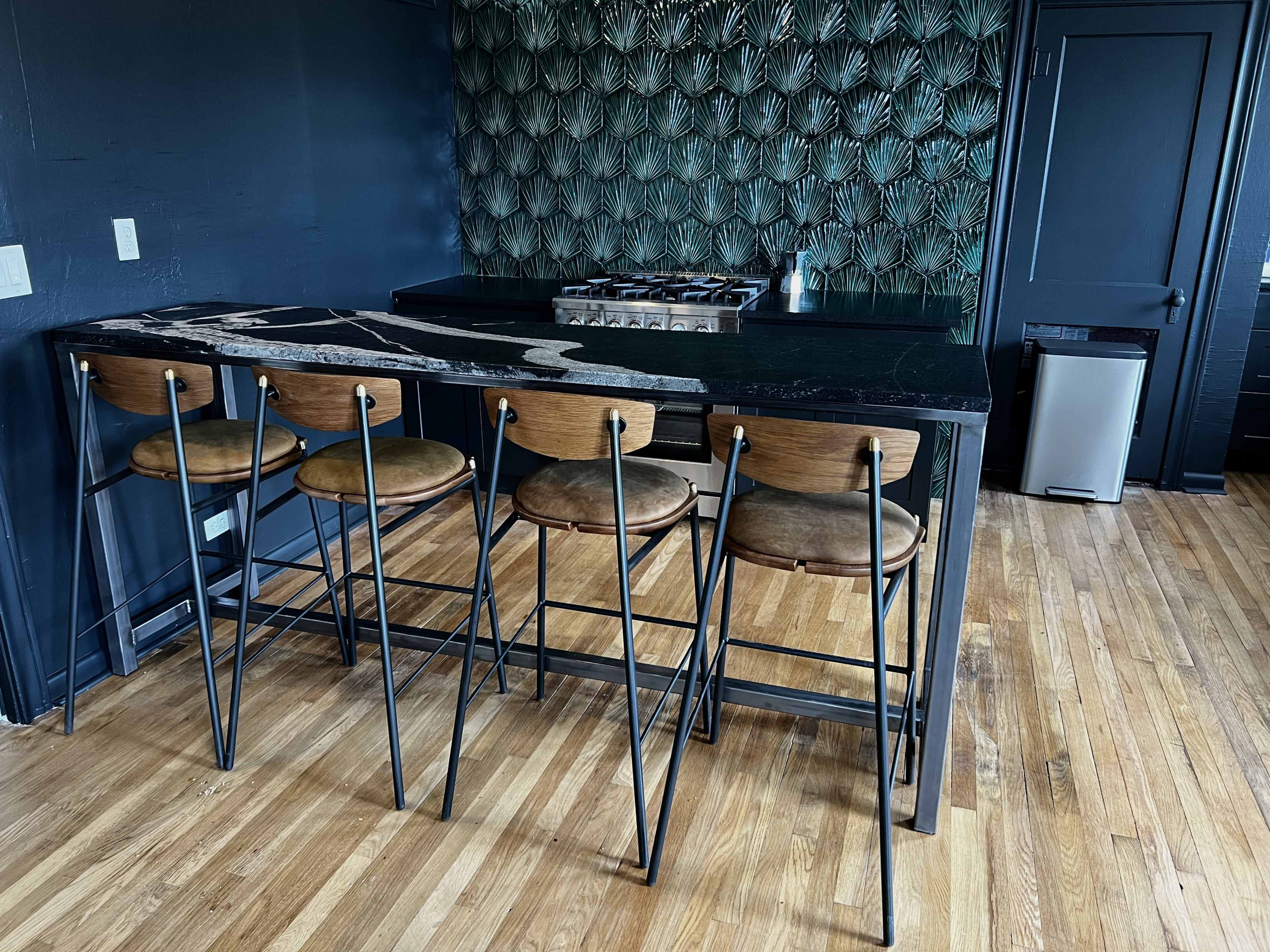 A modern kitchen features a bar with four wooden stools and a black countertop with a unique design, complemented by a textured green wall.