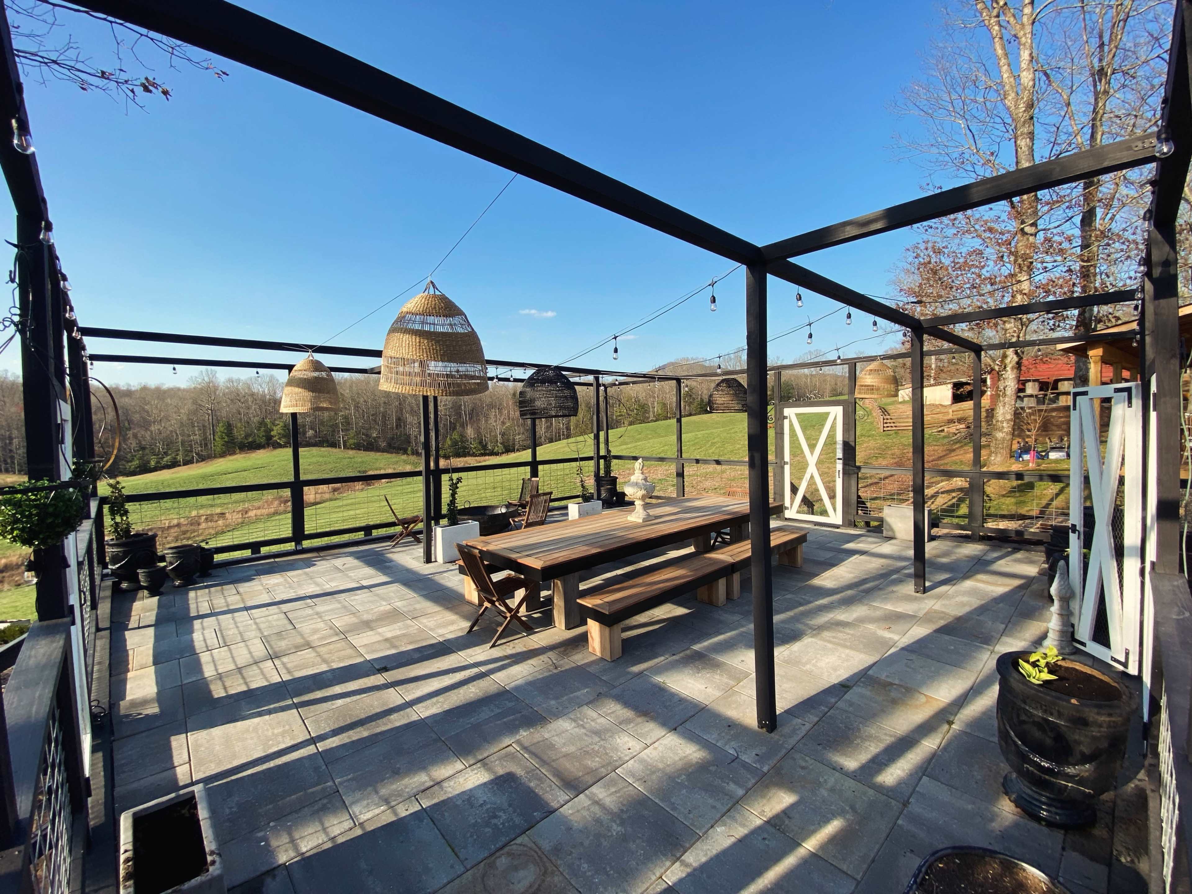 The image shows a spacious outdoor patio area with a large wooden dining table and hanging light fixtures, overlooking a green field and trees.